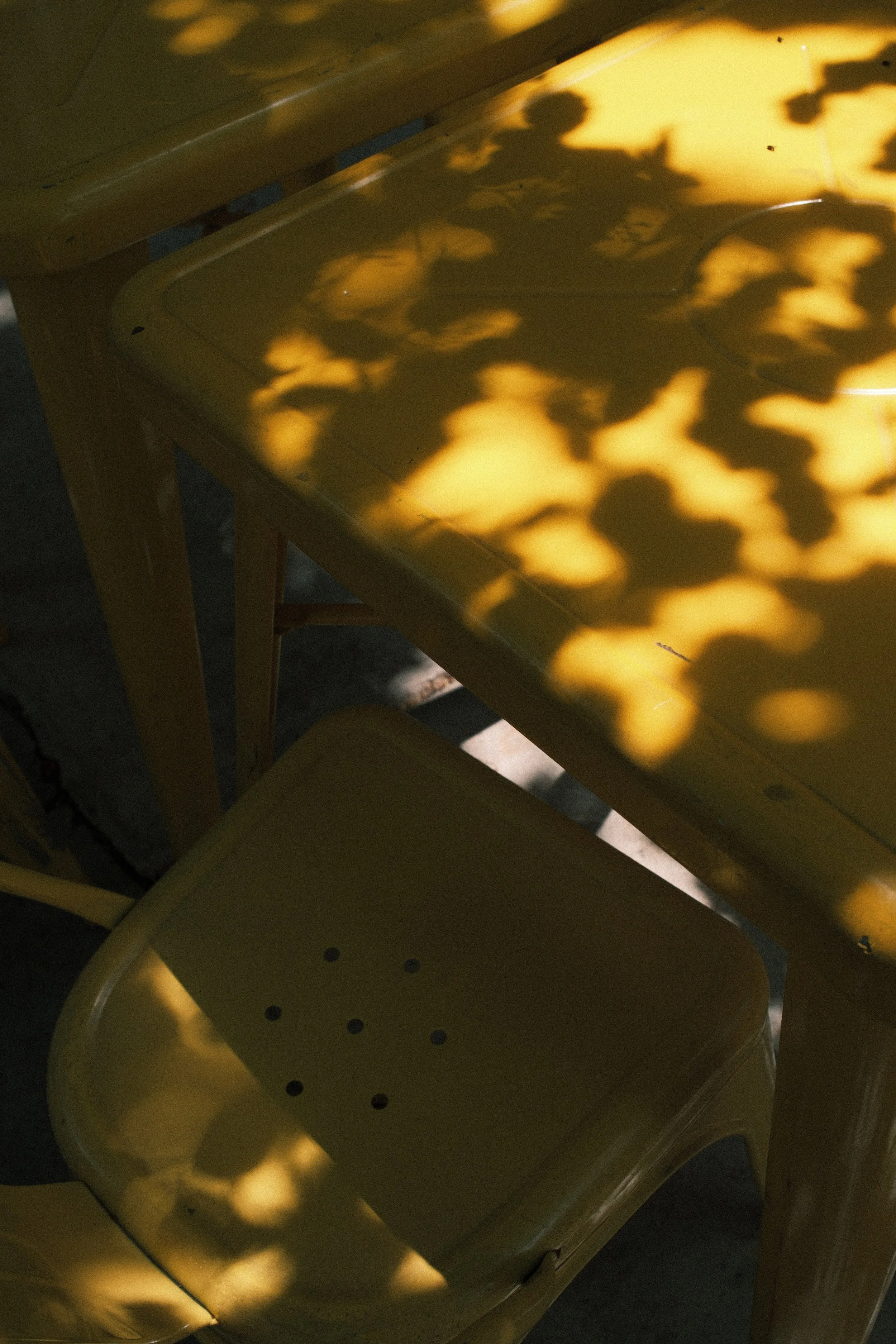 Yellow metal table with shadows of leaves and light patterns on its surface and a matching yellow chair underneath.