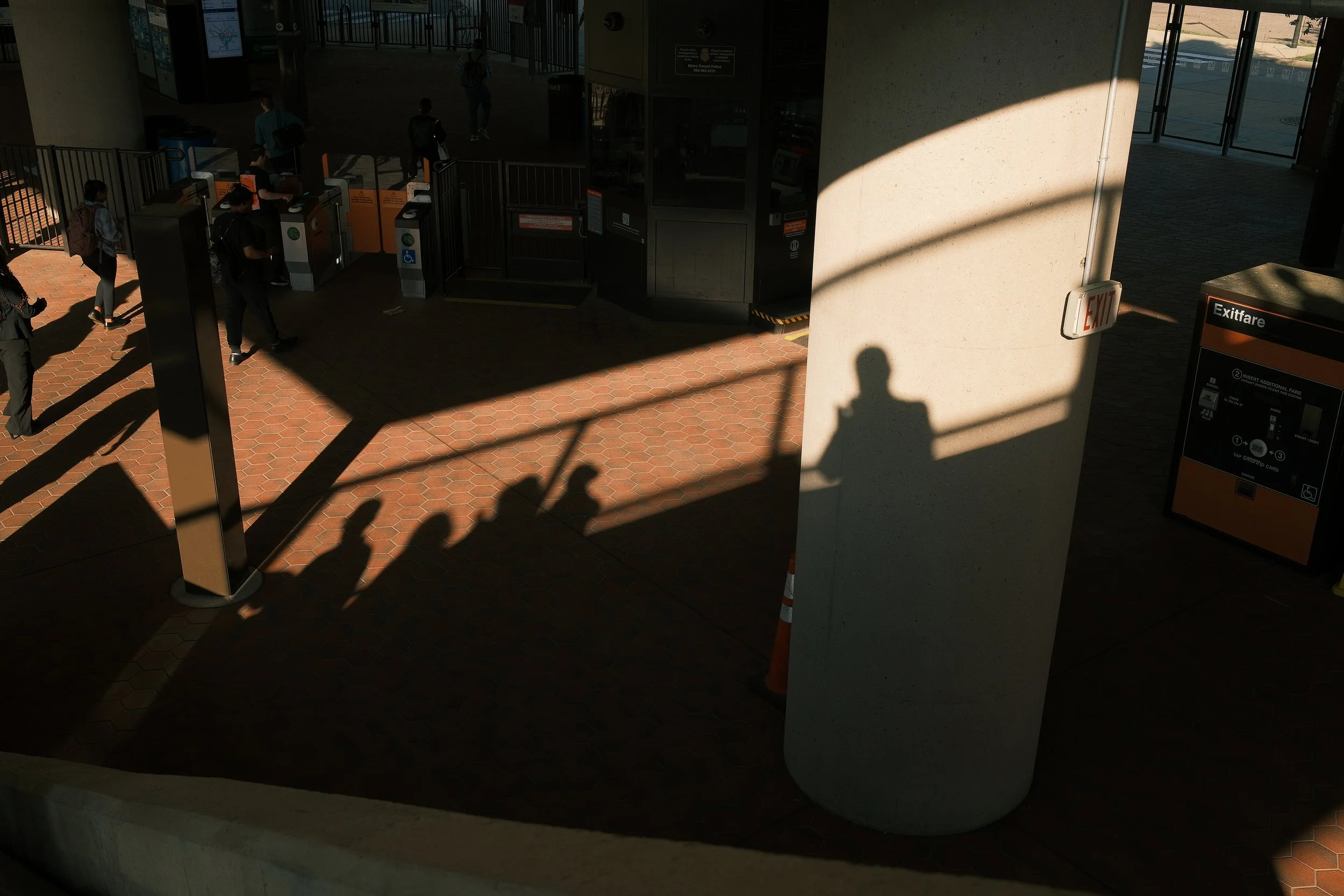 People walking through a covered outdoor area with ticket gates, a large concrete pillar with shadow, and an exit sign.