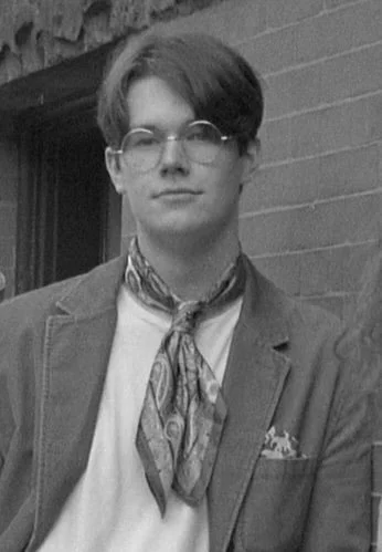 Photo of the artist: Connor Bechen. A young man wearing glasses, a blazer, and a patterned scarf around his neck, outdoors in front of a brick wall.