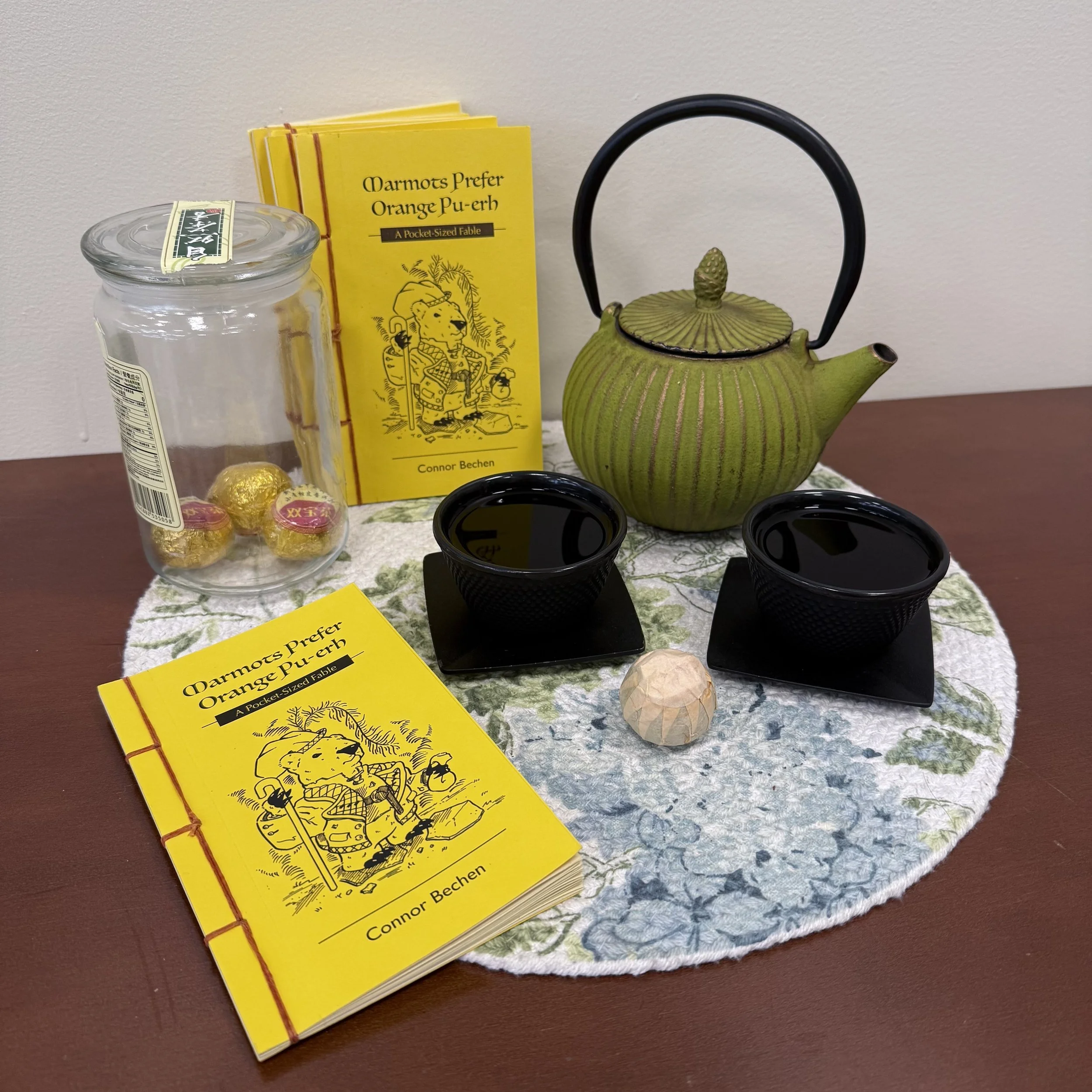 Tea setup with a green vintage teapot, two black teacups on black coasters, a jar containing wrapped tea oranges, a folded yellow book titled 'Marmots Prefer Orange Pu-erh' by Connor Bechen, and a white decorative cloth on a wooden table.