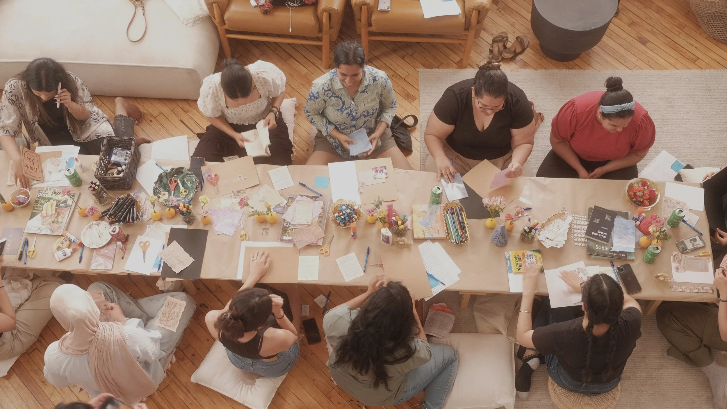 top-down view of a group of women gathered around a large table working on craft projects, with supplies like paper, scissors, and markers scattered across the table, in a cozy room with wooden floors and a beige couch.