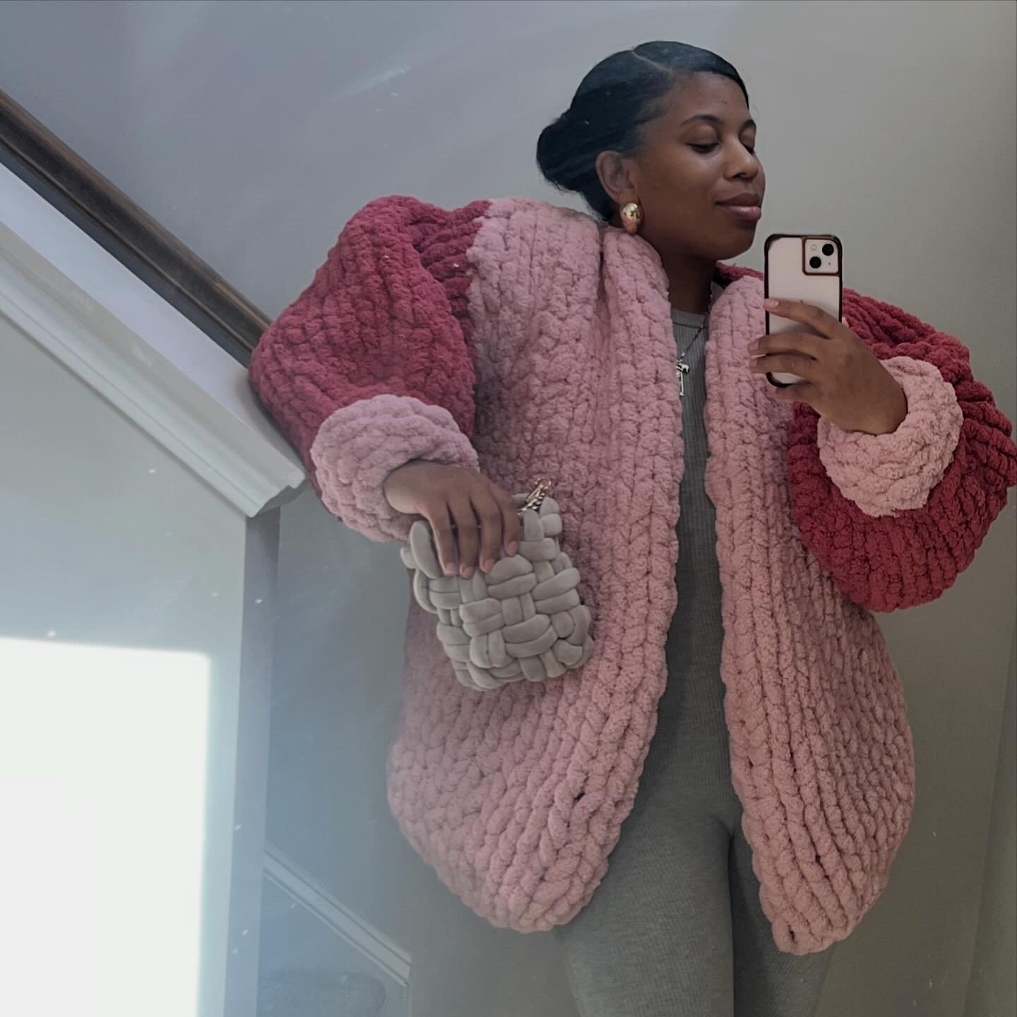 Woman taking a mirror selfie in a pink and red textured coat, holding a beige woven handbag, wearing earrings and keychain, against a neutral background.