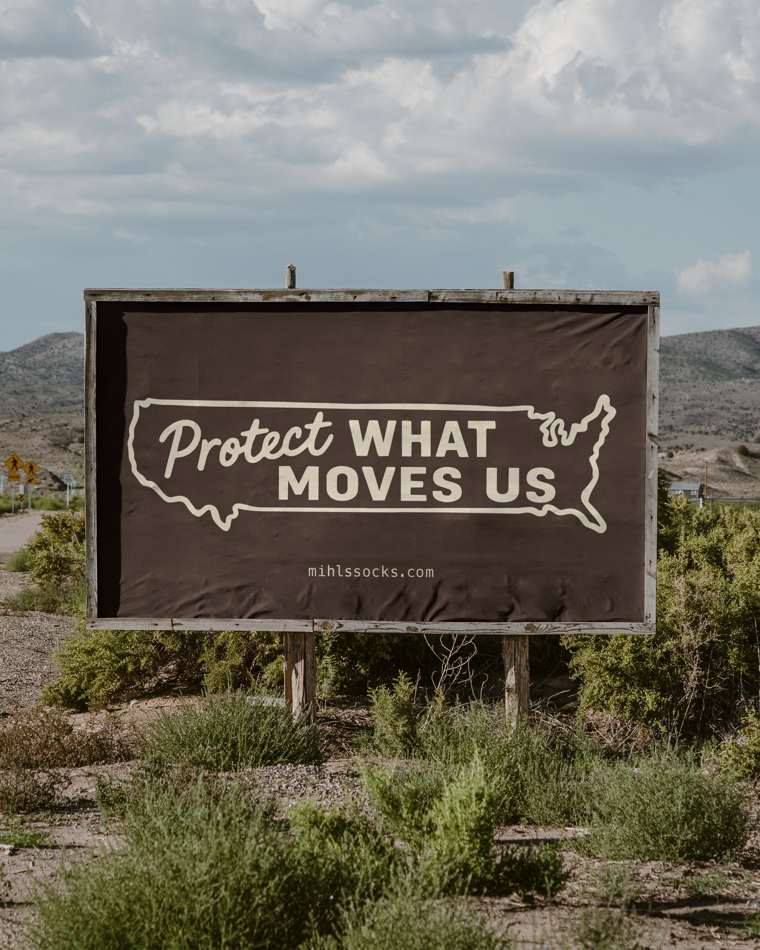 Billboard with the outline of the United States and text that reads 'Protect What Moves Us', website mihlssocks.com, in a desert landscape with bushes and hills under a partly cloudy sky.
