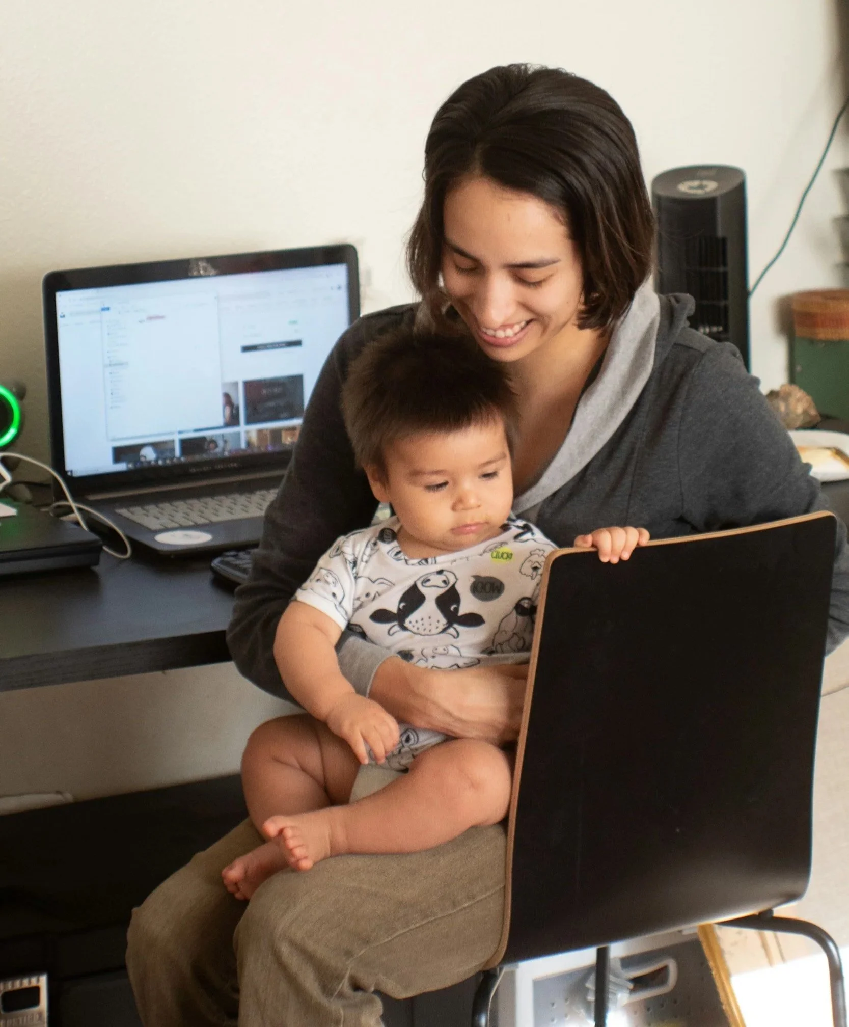 A young woman sitting on a chair with a small child on her lap, looking at a laptop screen. The woman is smiling, and the child is focused on the laptop, in a home office setting.