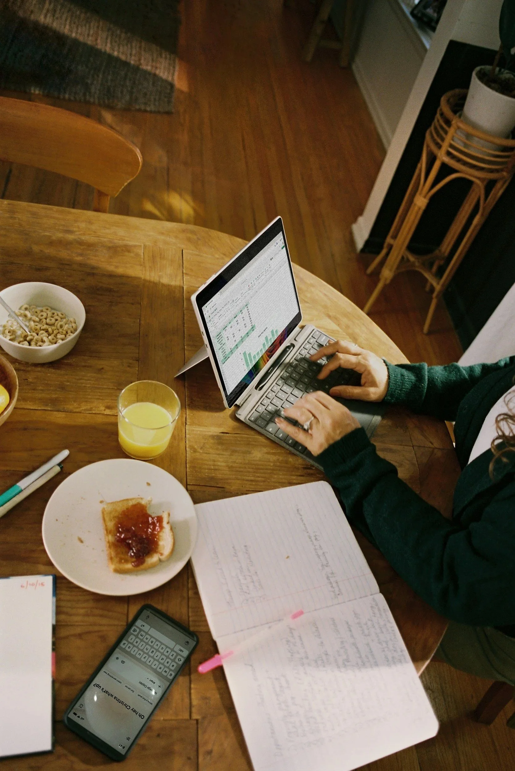 A person working on a laptop at a wooden dining table with a bowl of cereal, a glass of orange juice, a plate with toast and jam, a smartphone, a notebook, and pens.