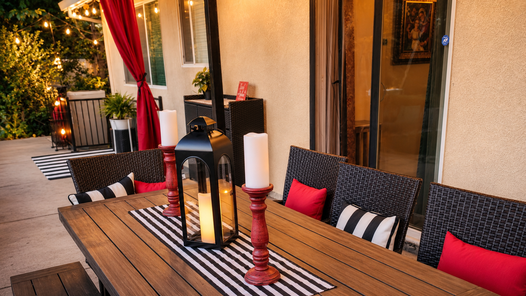 Budget-friendly backyard makeover with a cozy outdoor dining area, wooden patio table, black lantern centerpiece, red accents, striped pillows, string lights, and warm evening patio decor.