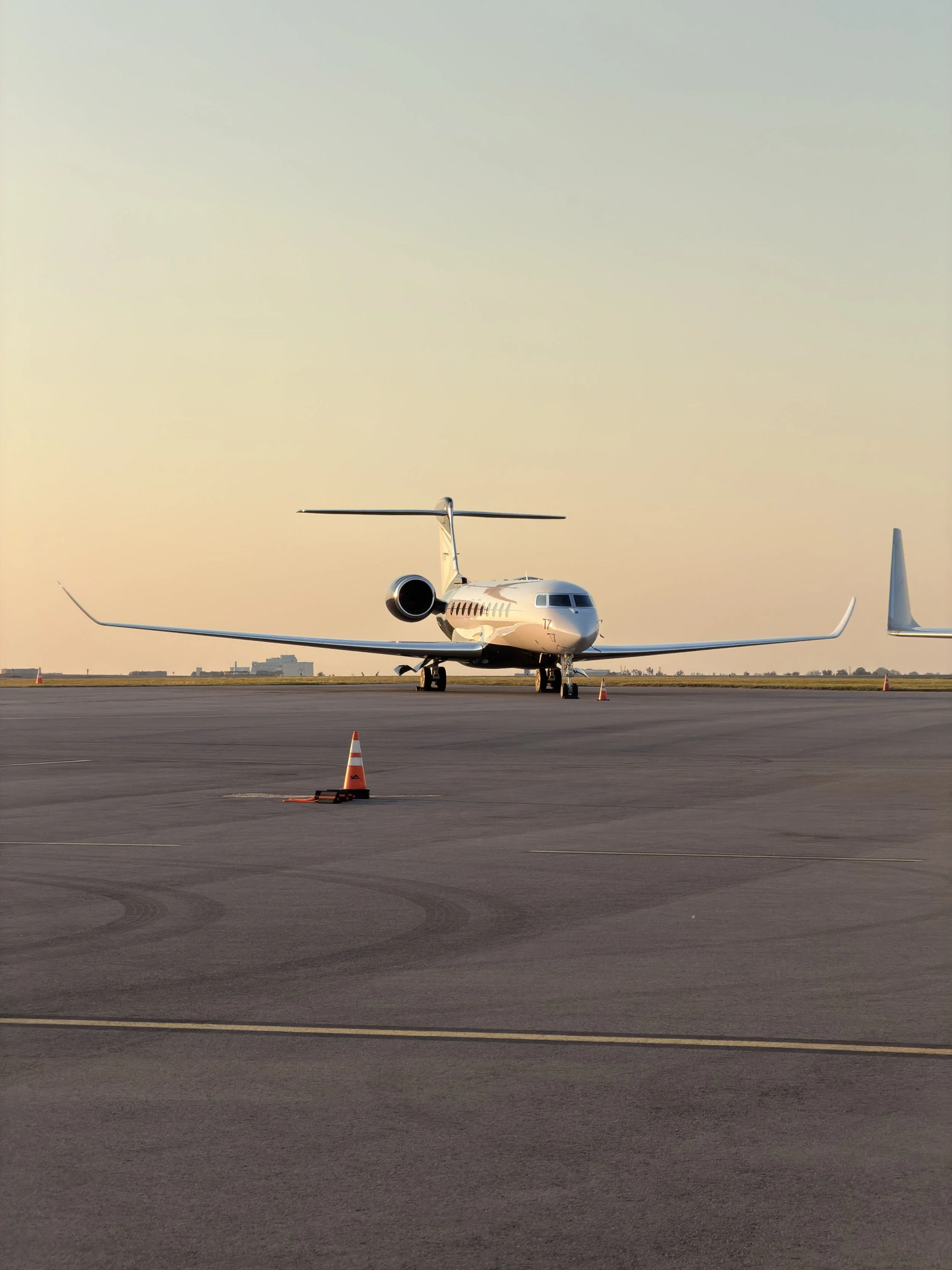 A white private jet parked on an airport tarmac with orange traffic cones and a city skyline in the background during sunset.
