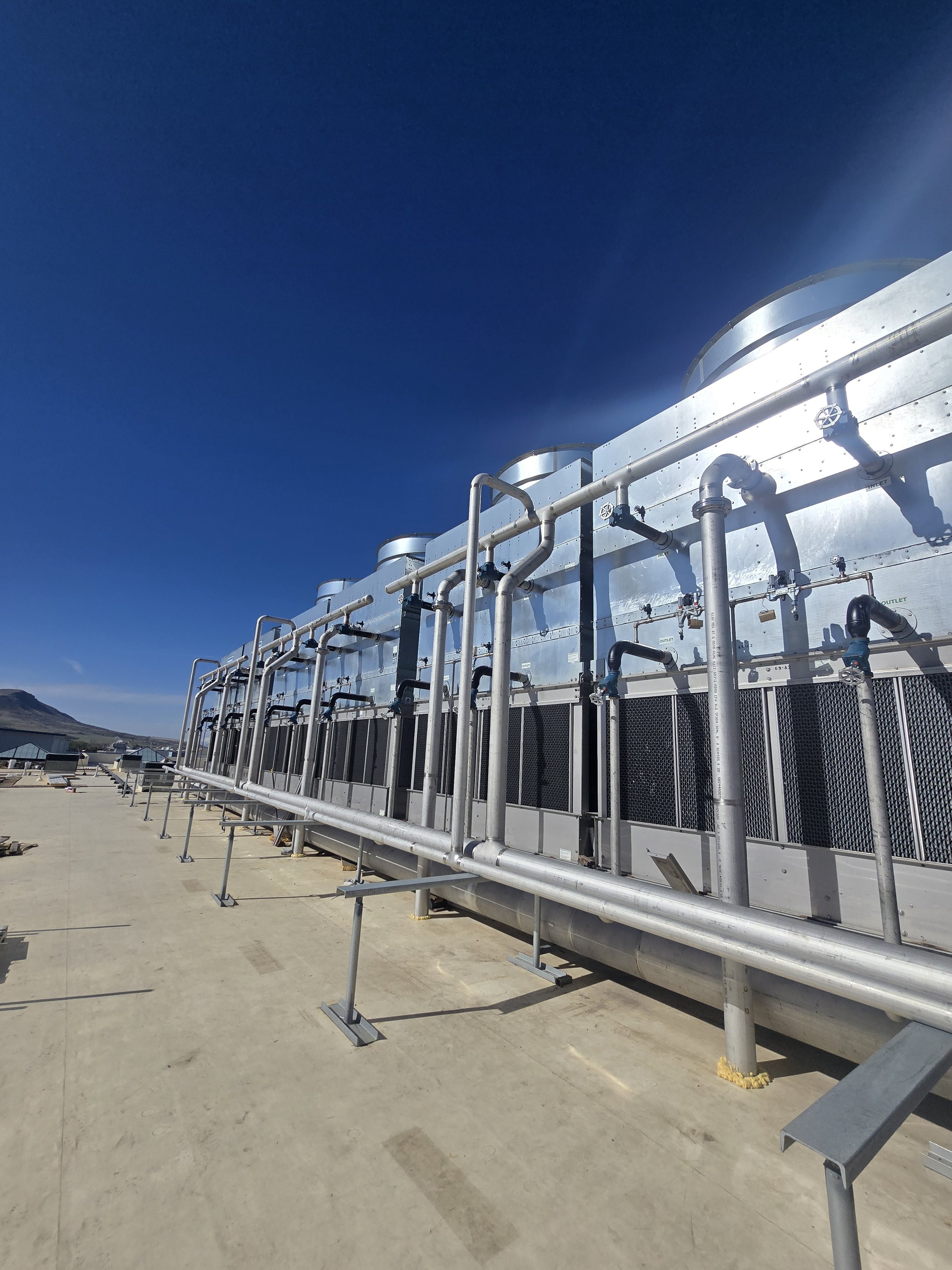 Industrial rooftop with large metal cooling or HVAC units, pipes, and vents against a clear blue sky, mountains visible in the background.