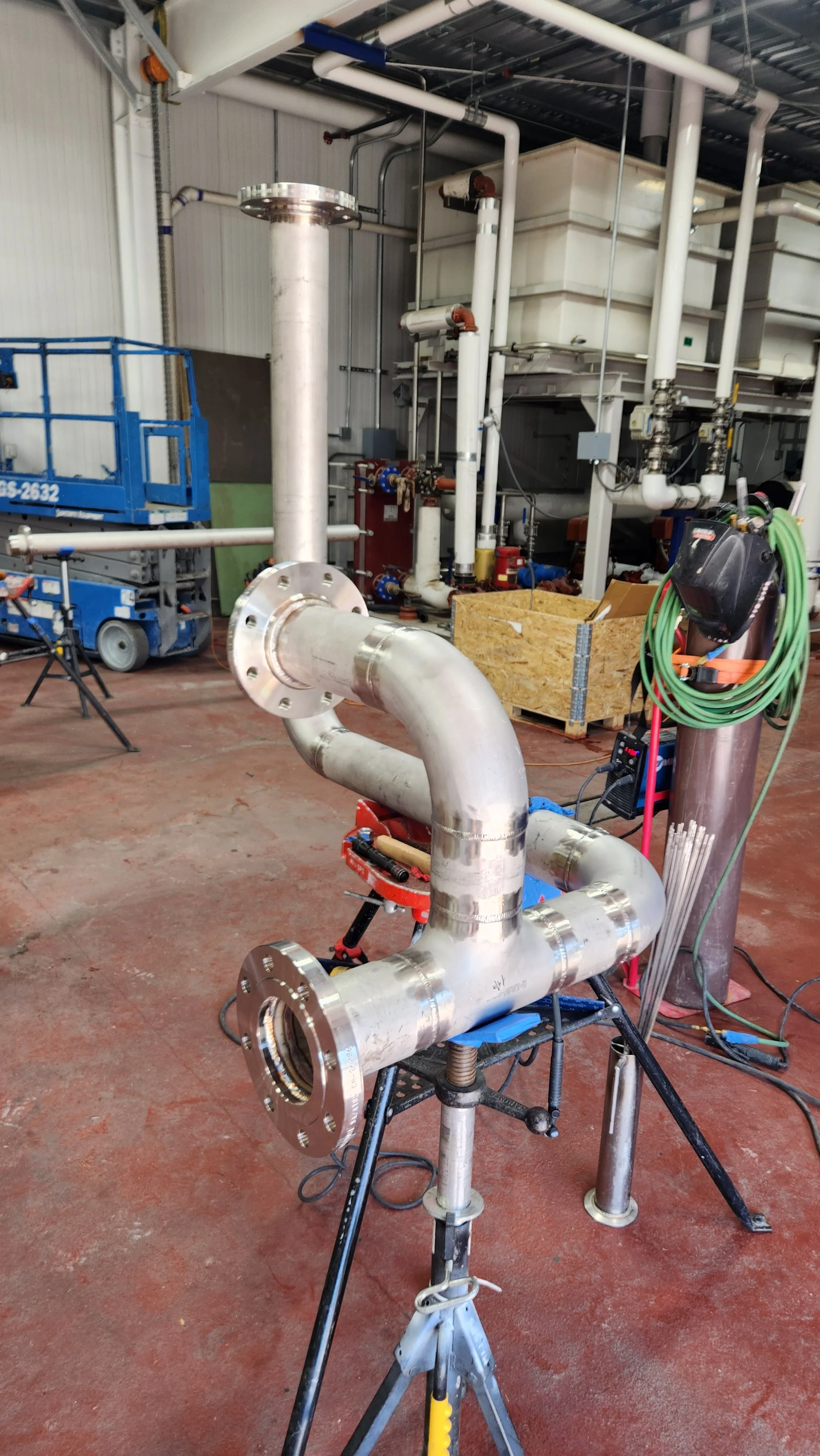 Industrial piping setup with large metal pipes, valves, hoses, and equipment in a warehouse or factory setting.