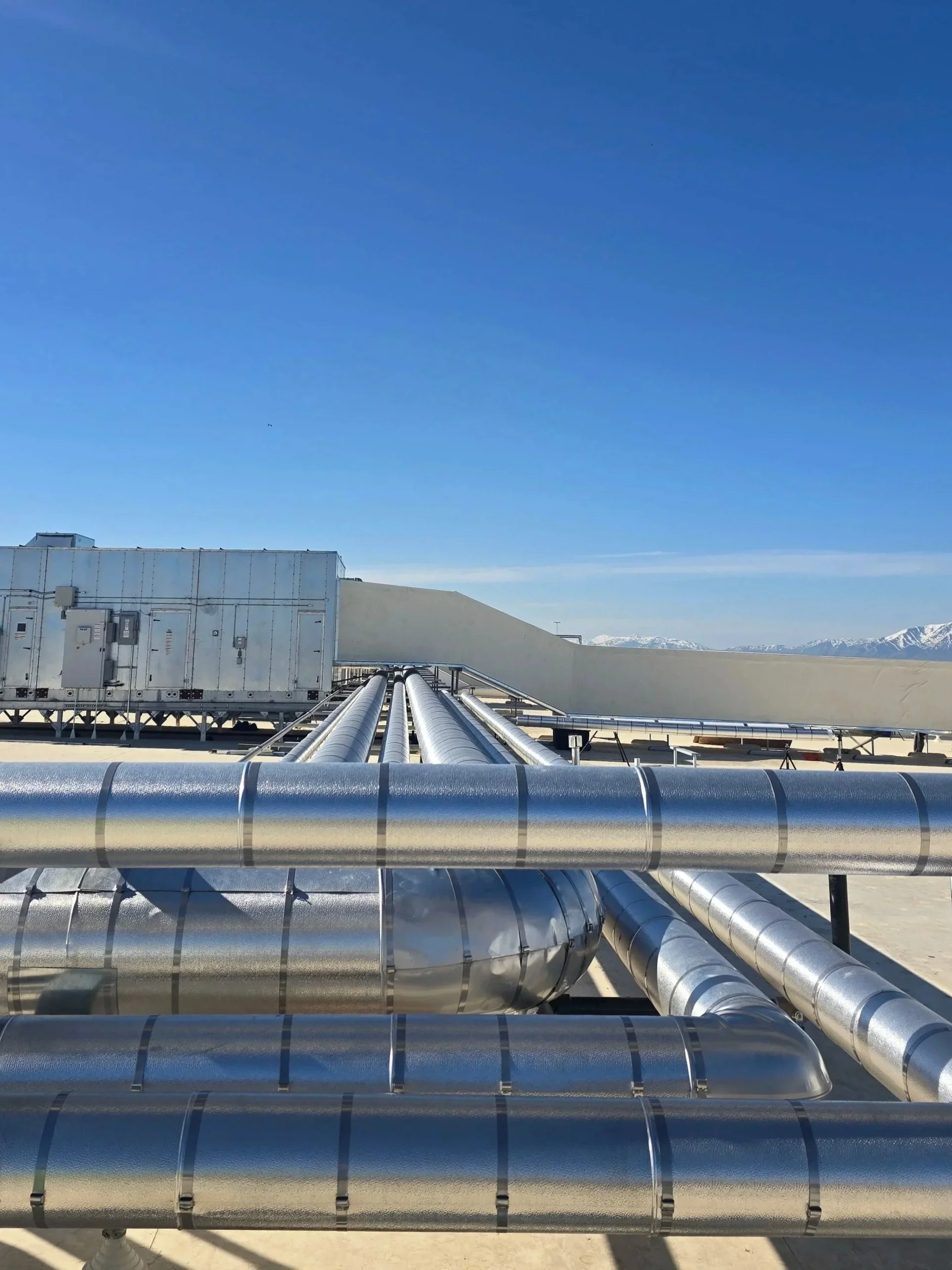 Multiple large, shiny, metallic pipes running parallel on a flat rooftop against a clear blue sky with distant snow-capped mountains.