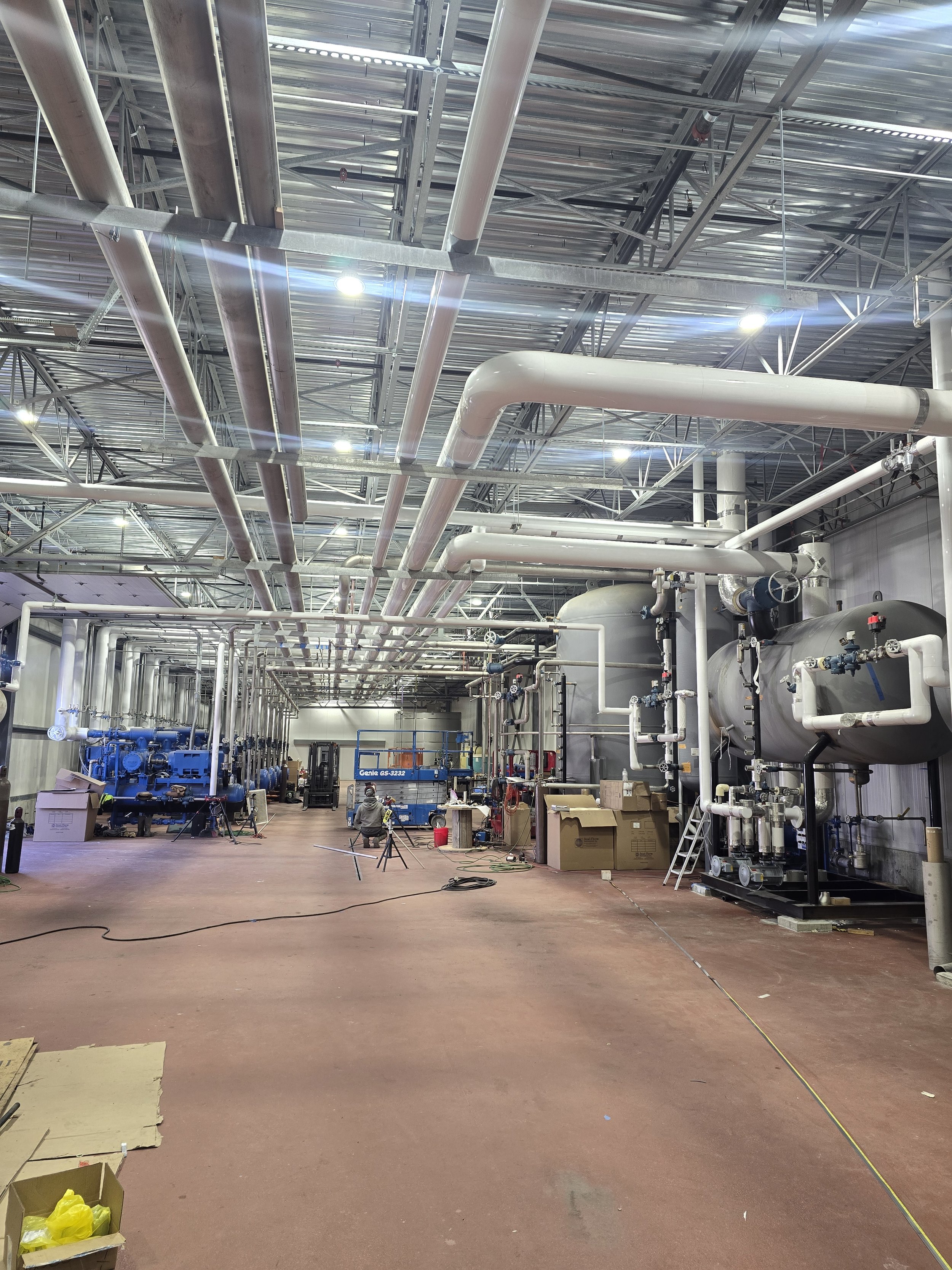 Industrial facility or warehouse with exposed ceiling pipes and equipment, some workers, boxes, and machinery scattered on the floor.
