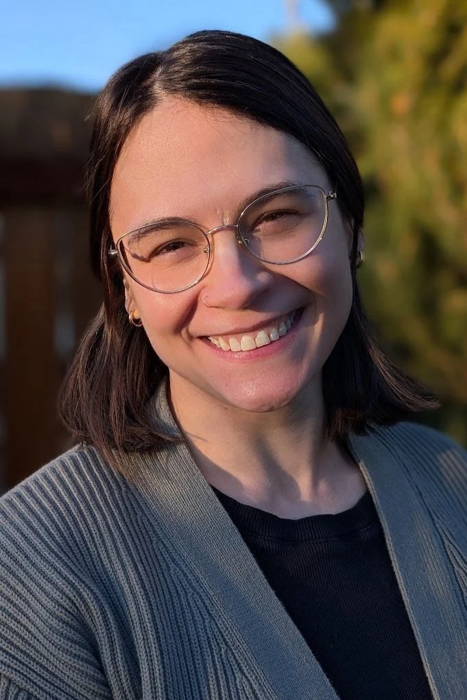 A woman with glasses smiling outdoors with trees and a building in the background.