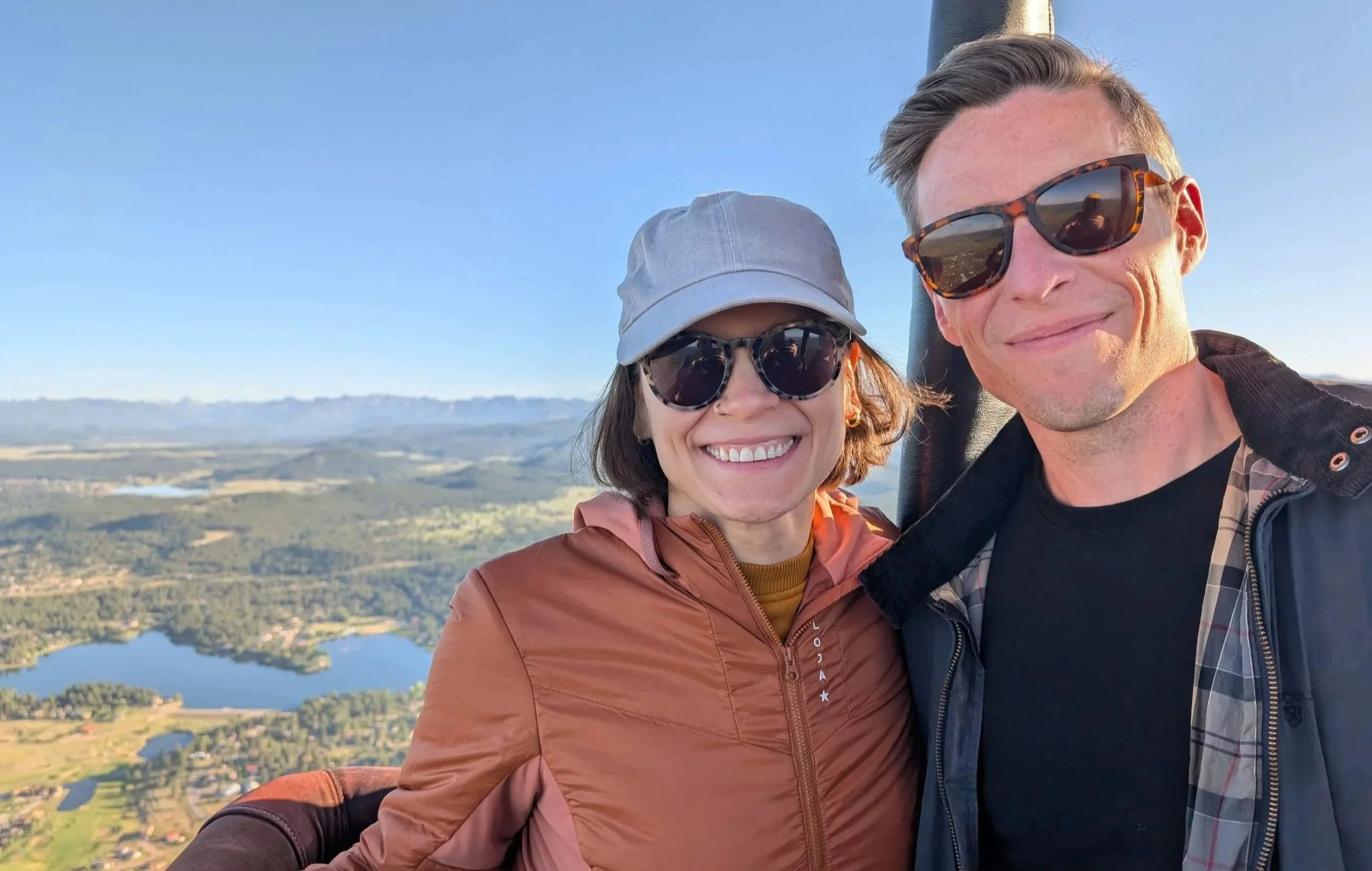 Smiling couple wearing sunglasses and casual outdoor clothing taking a selfie with a scenic landscape of lakes, fields, and mountains in the background.