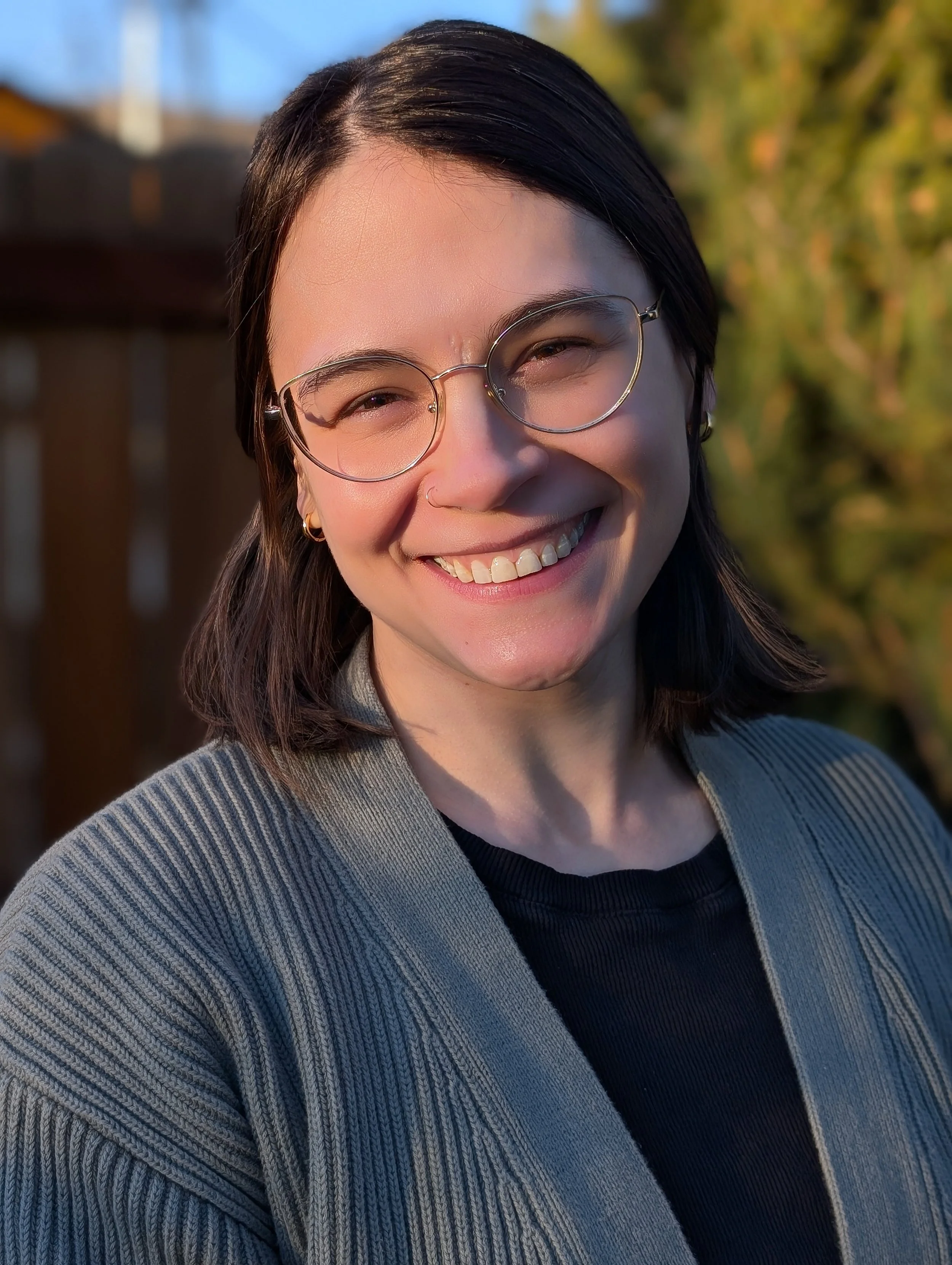 A young woman with glasses, dark brown hair, and a nose ring smiling outdoors during daylight. She is wearing a striped jacket over a black shirt, with trees in the background.