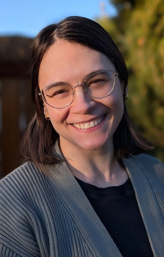 Close-up of a smiling woman with shoulder-length dark hair, wearing round glasses, a black top, and a gray blazer outdoors with green foliage and a blue sky in the background.