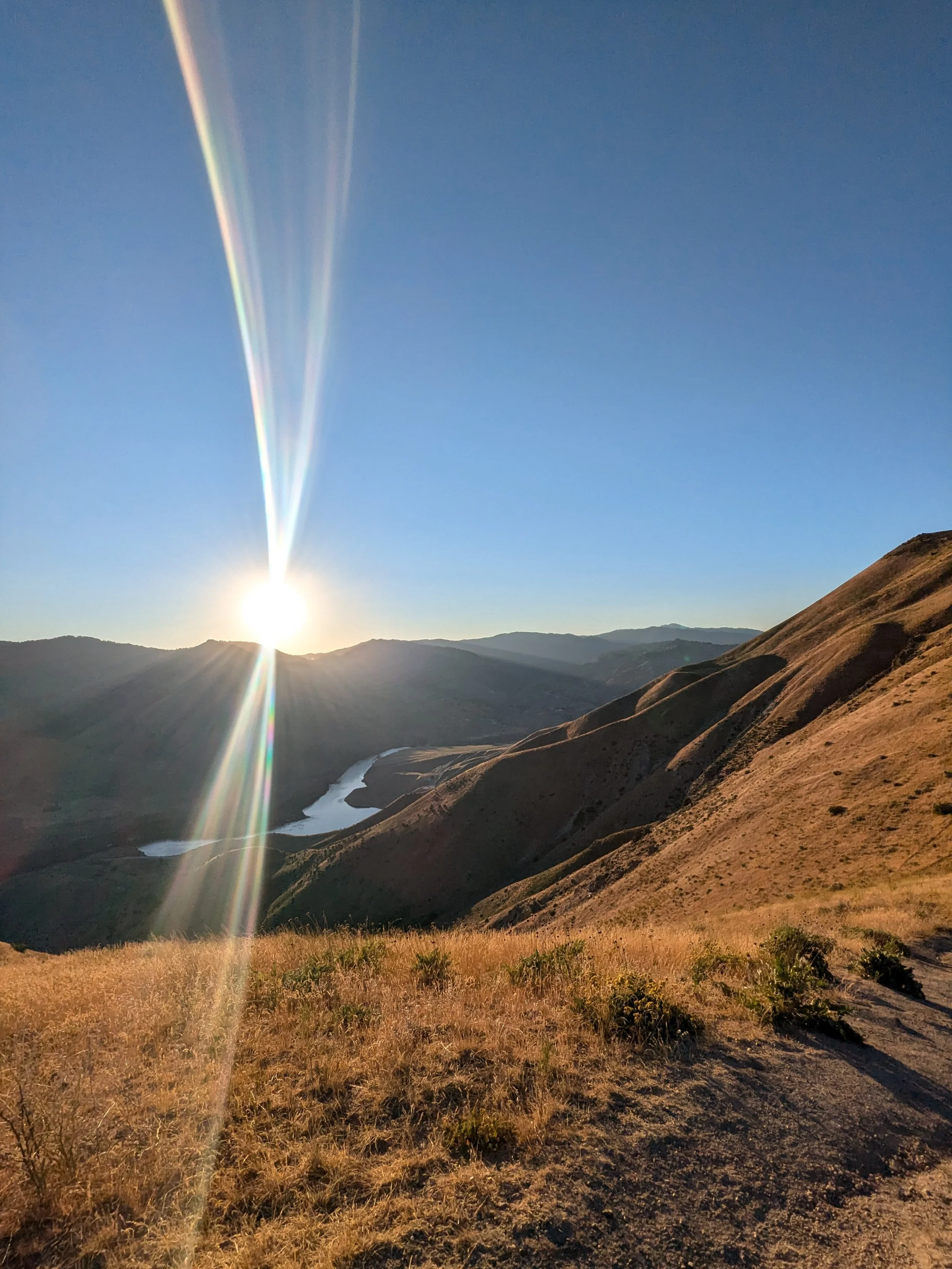 Sunset over hilly landscape with a winding river, dry grass, and mountain ranges in the distance.