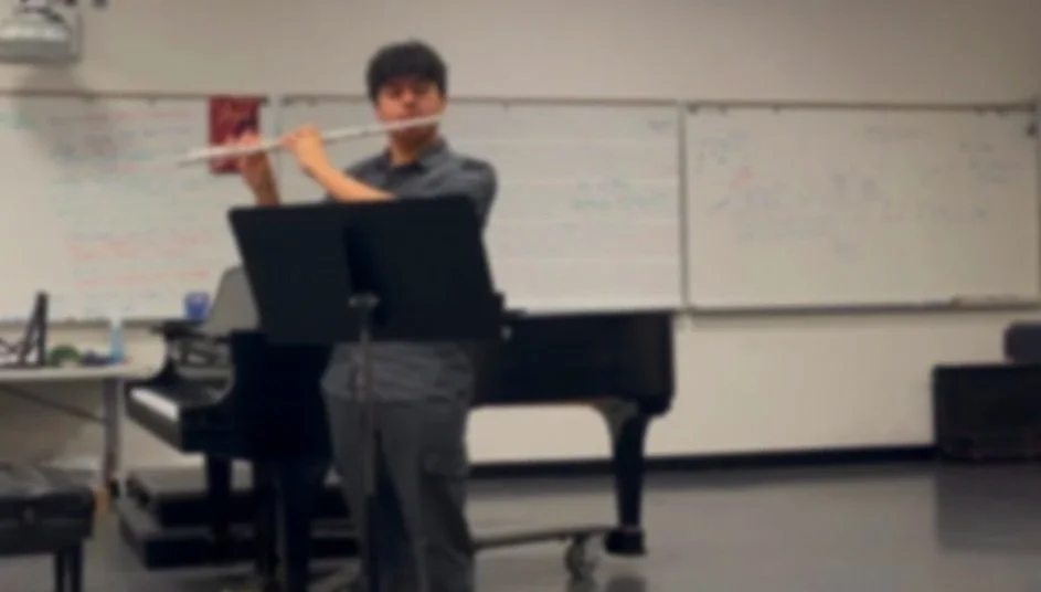 A person playing the flute in a classroom with a whiteboard and piano in the background.
