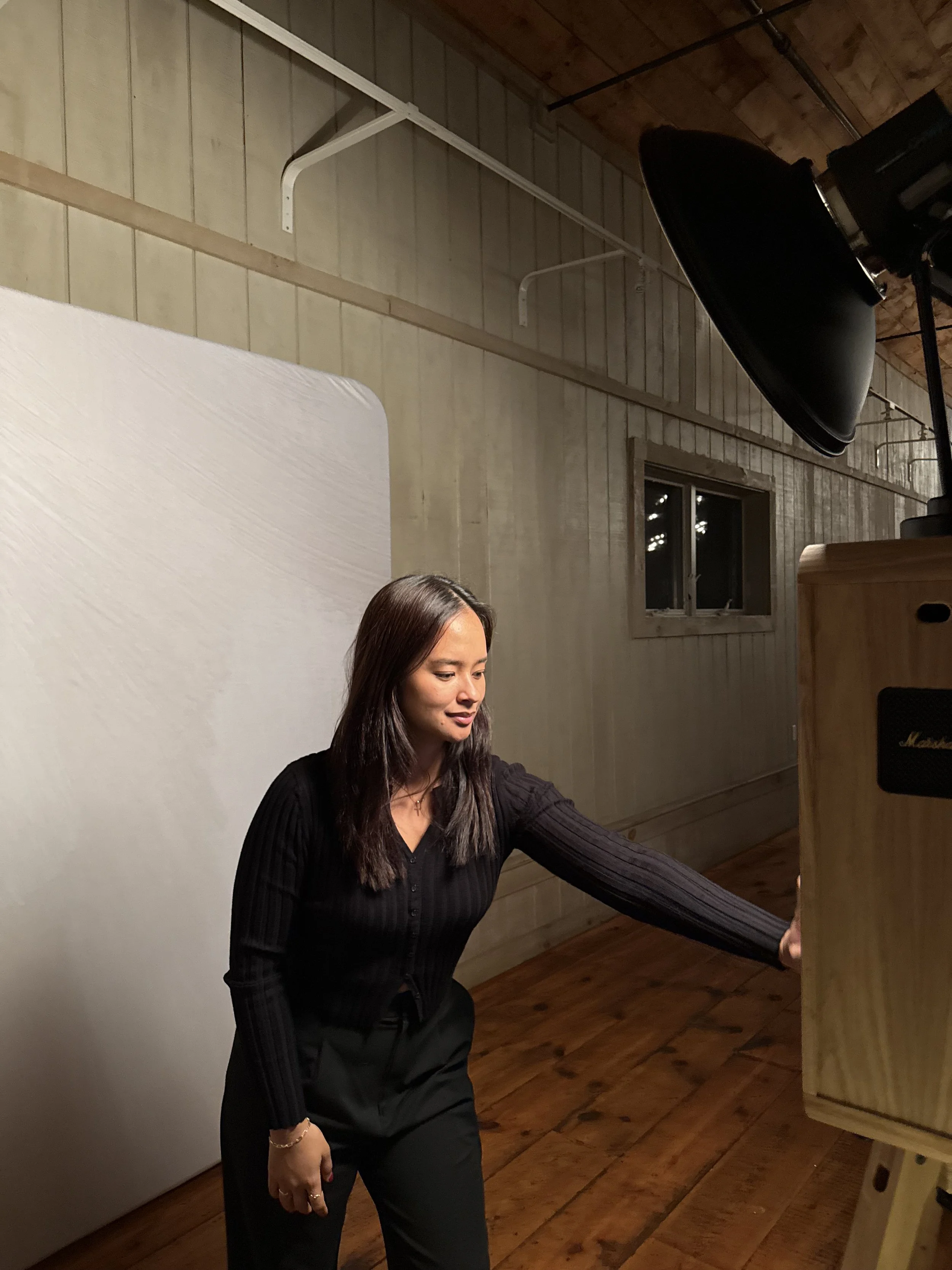 A woman with long dark hair wearing a black long sleeve top and black pants, standing inside a wooden room with a white wall on her left, a small window behind her, and a black studio lamp overhead.