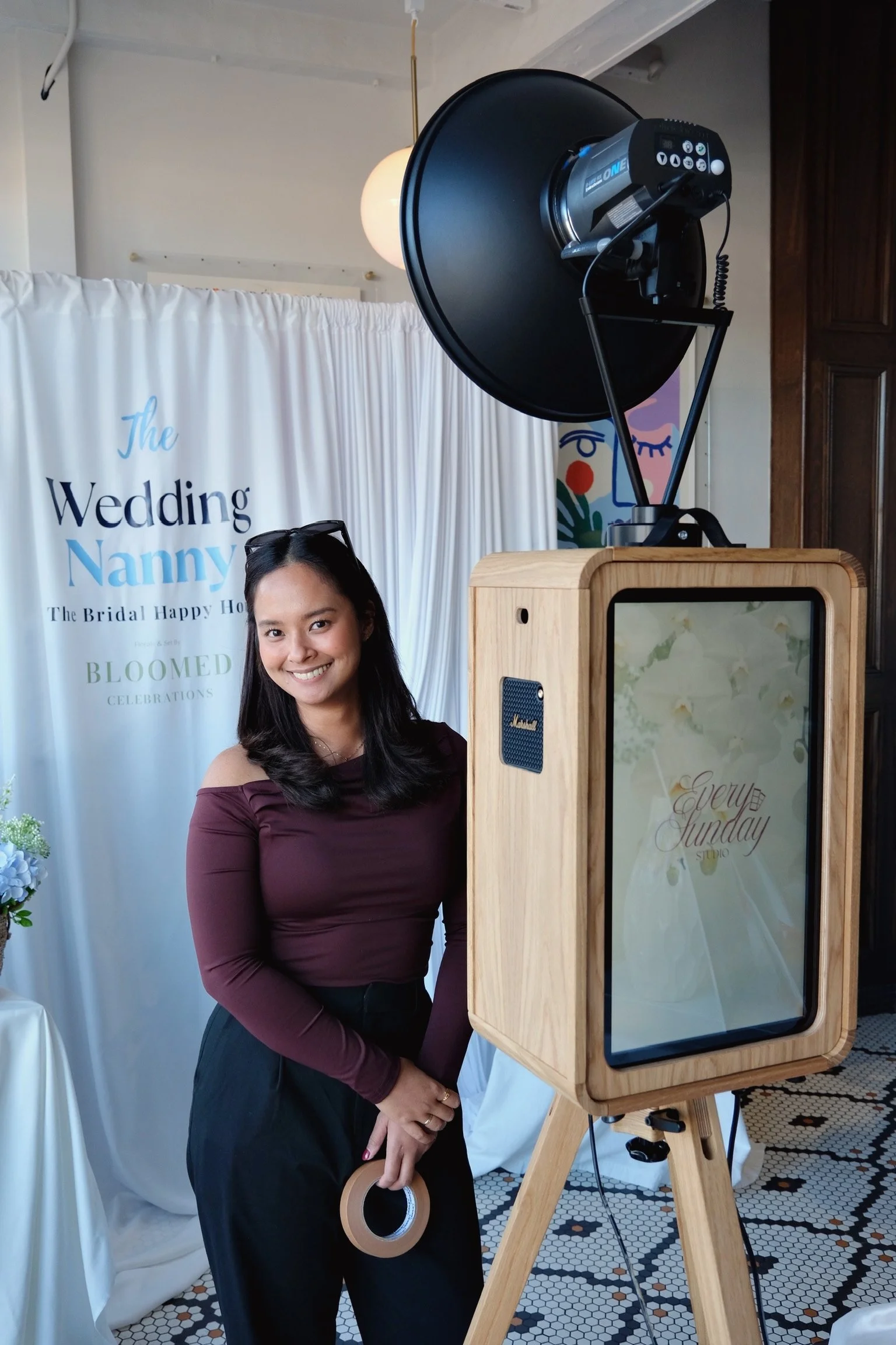 A woman in a wine-colored off-shoulder top smiling at a photo booth with a sign behind her reading 'The Wedding Nanny'. She is holding a roll of tape and standing next to a wooden photo booth with a sign that says 'Every Sunday'. There is professional lighting equipment above her, and the setting appears to be at a wedding celebration with a patterned black-and-white tiled floor.