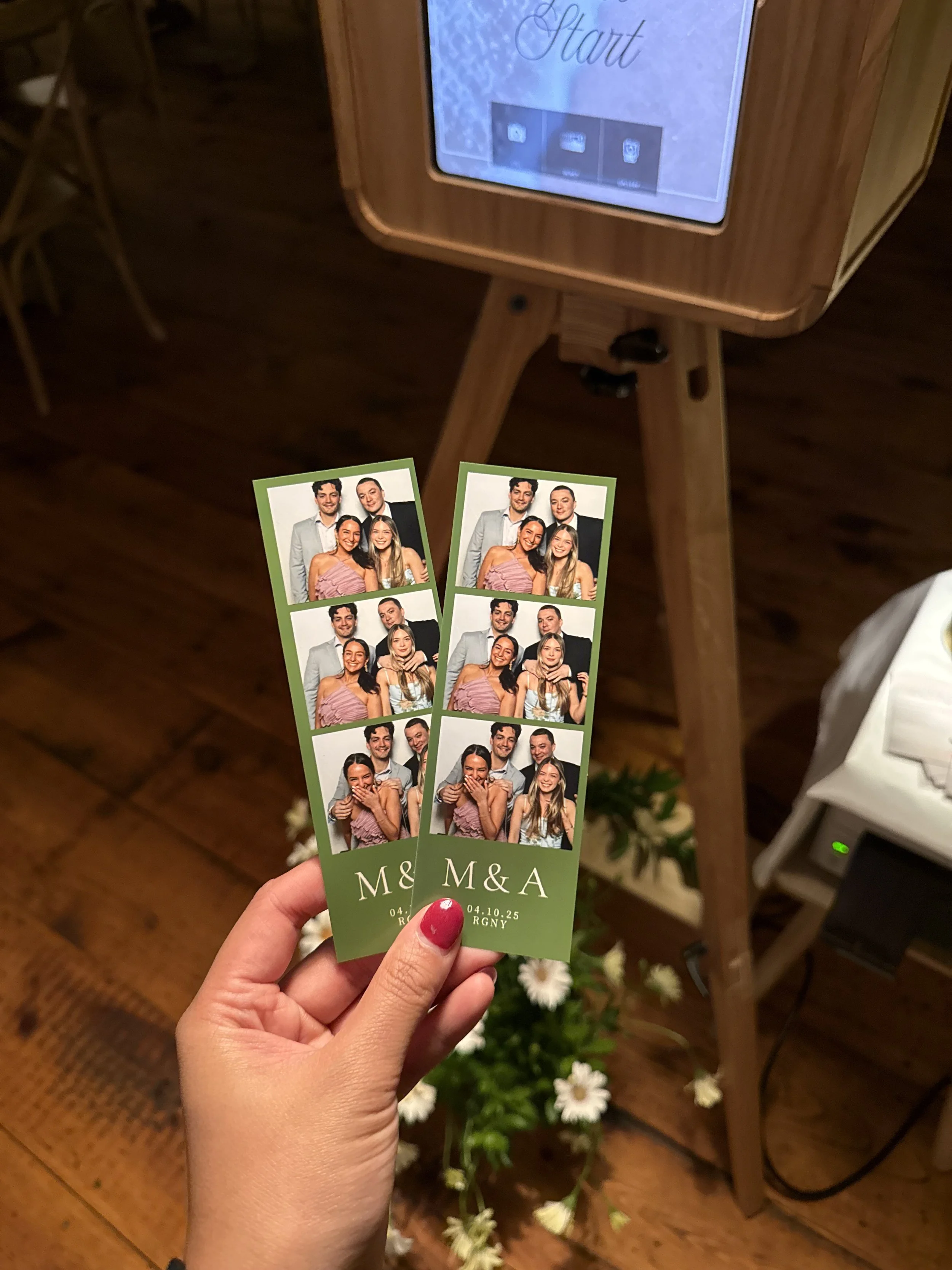 Two photo booth tickets held in a person's hand, with a wooden table and a photo booth screen displaying the word 'Start' in the background.