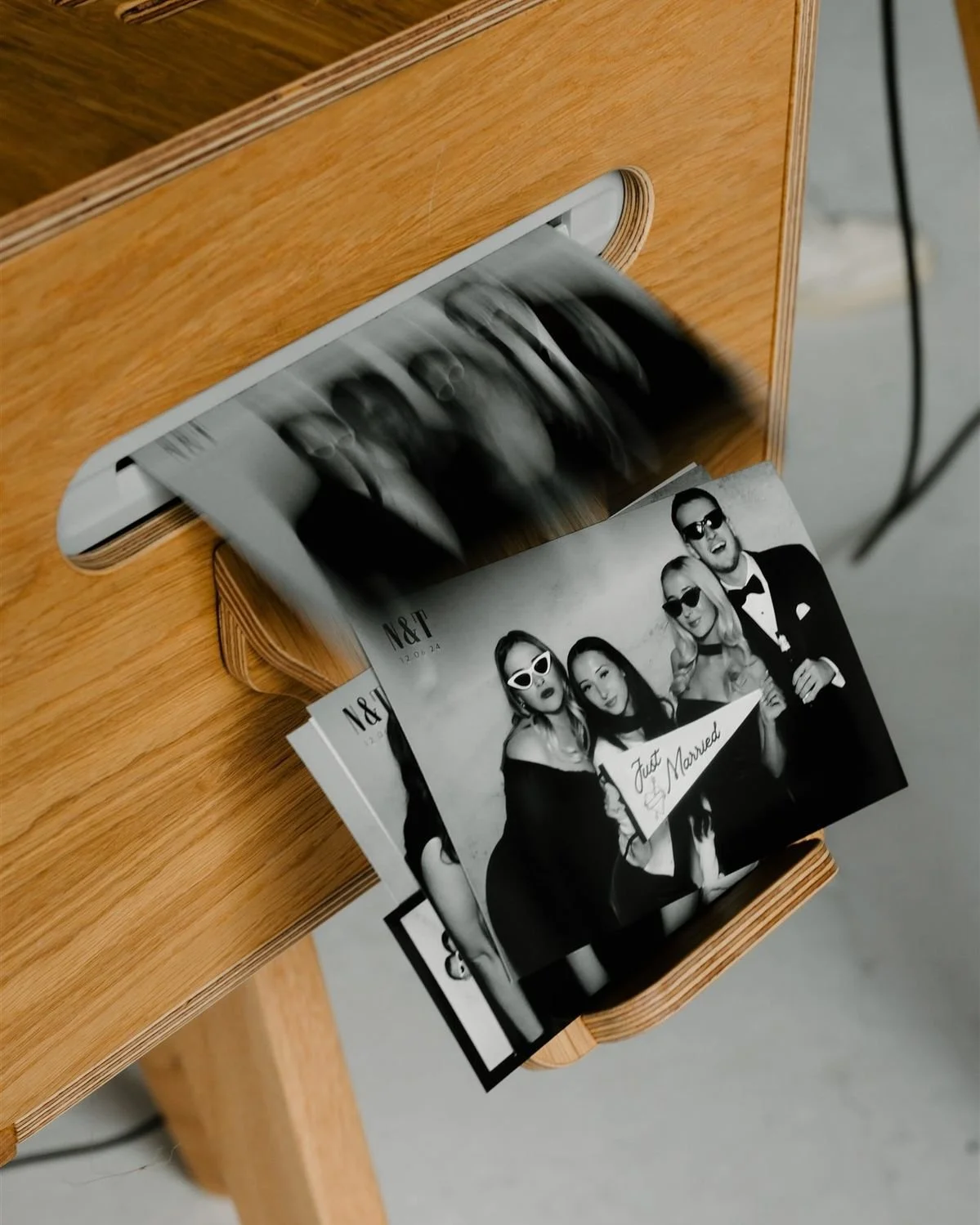 Photostrip being ejected from a photo booth, showing a black-and-white image of four people in formal attire, holding a sign that reads "Just Married," with some wearing sunglasses.