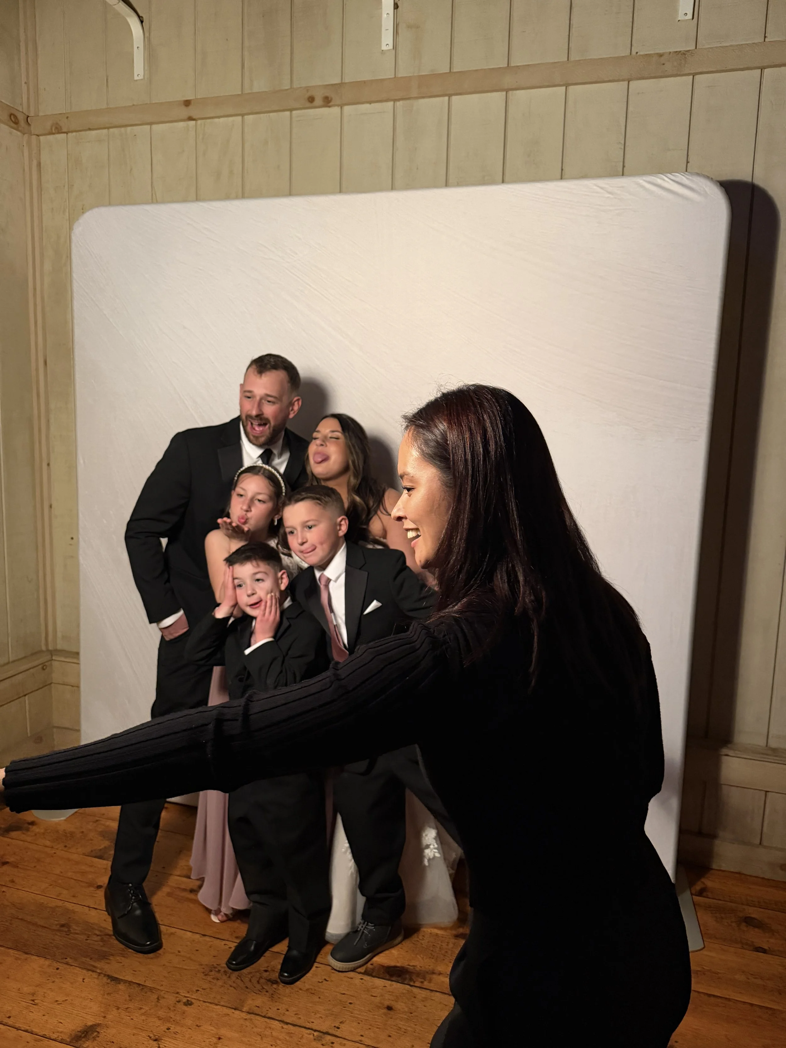 A woman is taking a group photo of a family in front of a white backdrop in a wooden room, with seven people making funny faces and poses.