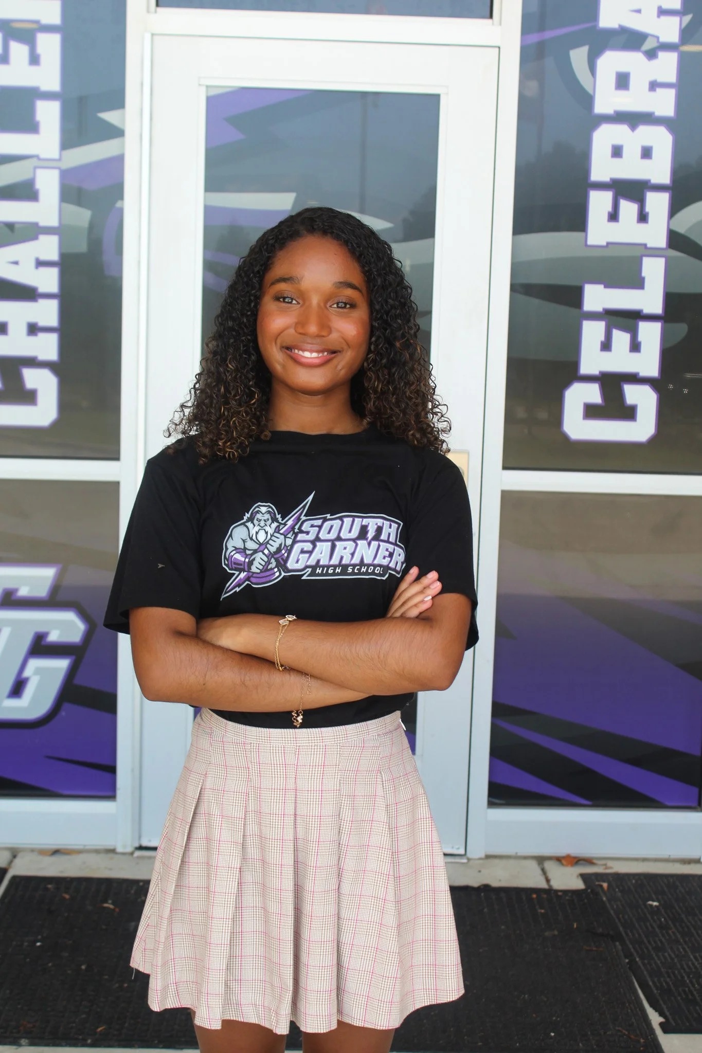 Young woman standing with arms crossed in front of school entrance, wearing a black T-shirt with 'South Garner High School' logo and a plaid skirt.