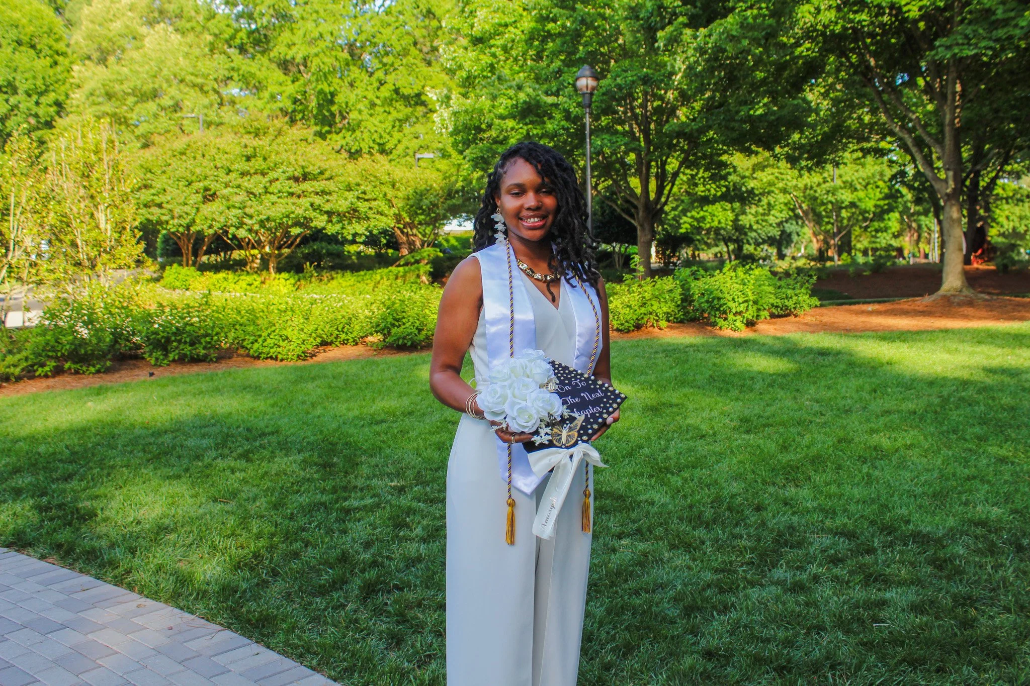 A woman in a white pantsuit holding a floral graduation cap pose outdoors on a grassy area with trees in the background.