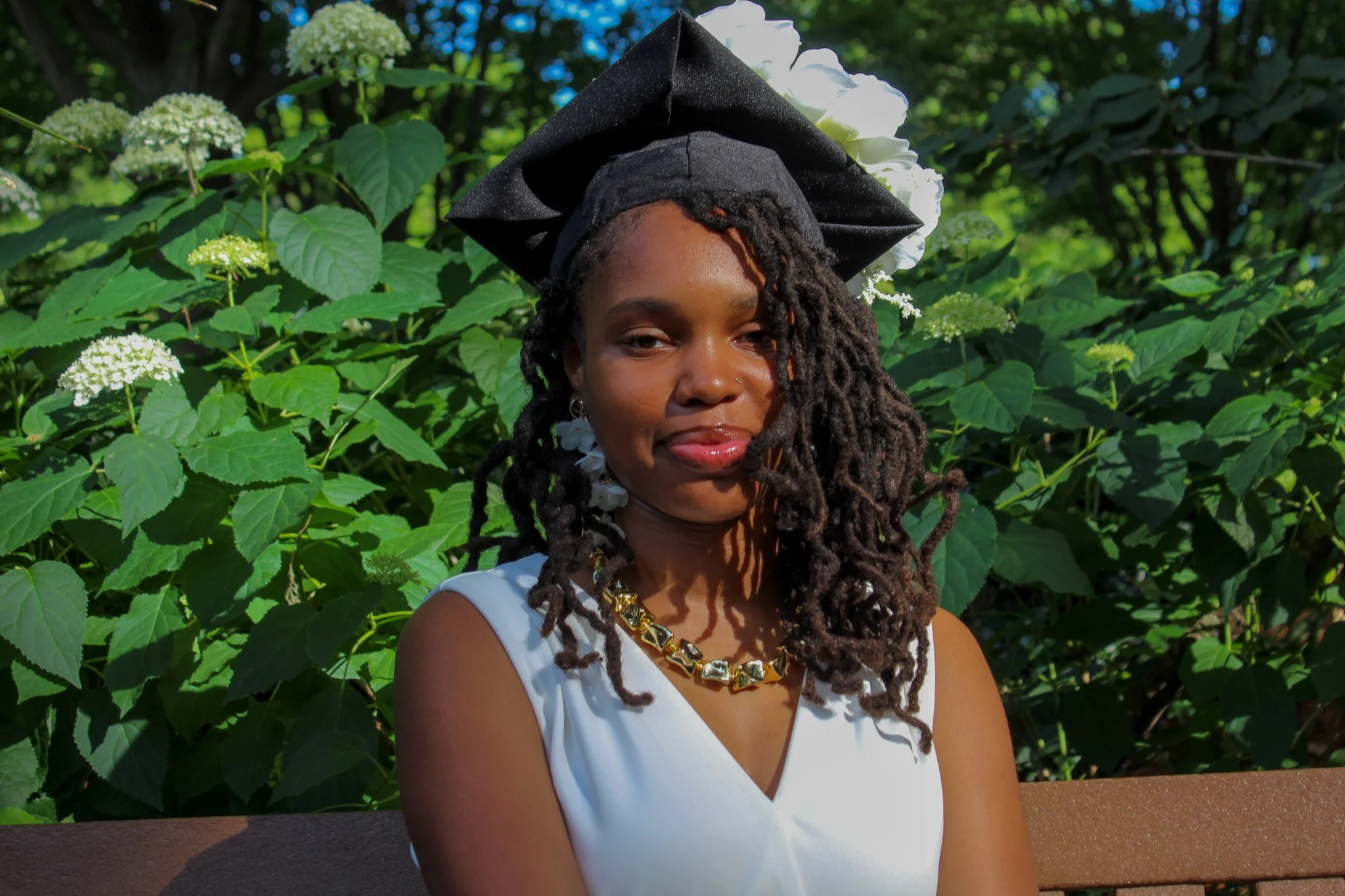 A woman with locs wearing a graduation cap and white pantsuit, sitting on a bench in front of green foliage with white flowers.