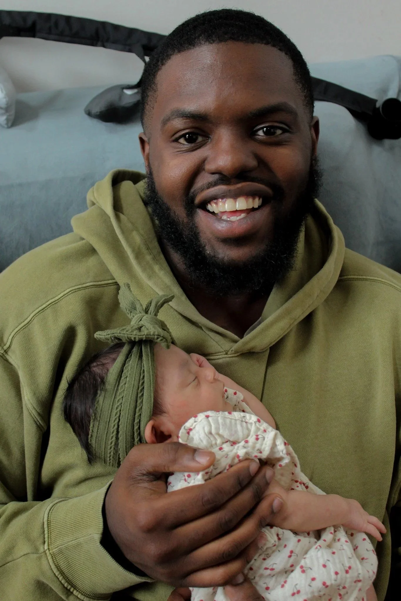 A smiling man with a beard holding a newborn baby wrapped in a white cloth with red dots, inside a room.
