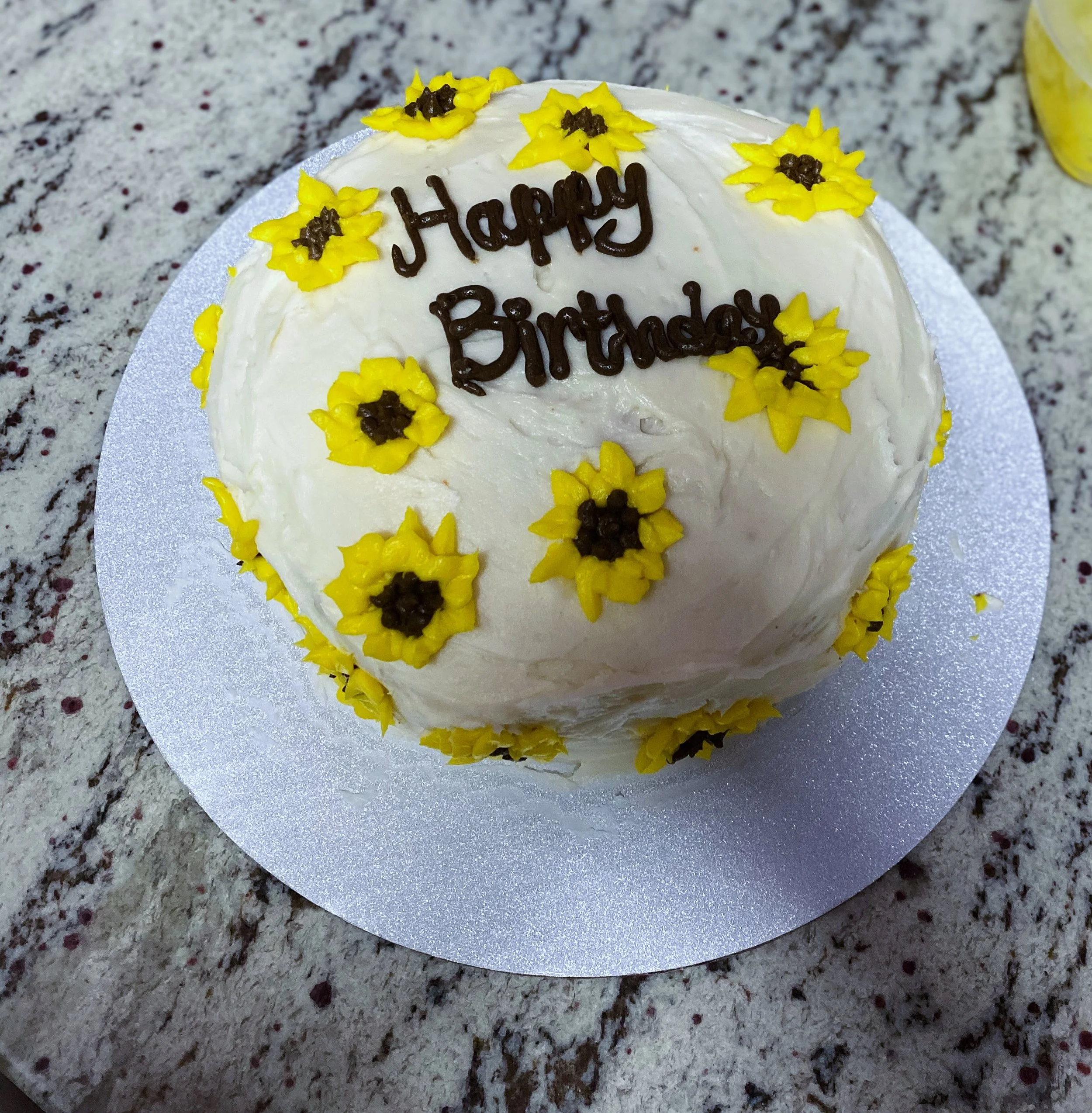 A round birthday cake with white frosting, decorated with yellow sunflower-shaped flowers with brown centers, and the words 'Happy Birthday' written in brown icing on top. The cake is placed on a silver cake board on a speckled granite countertop.