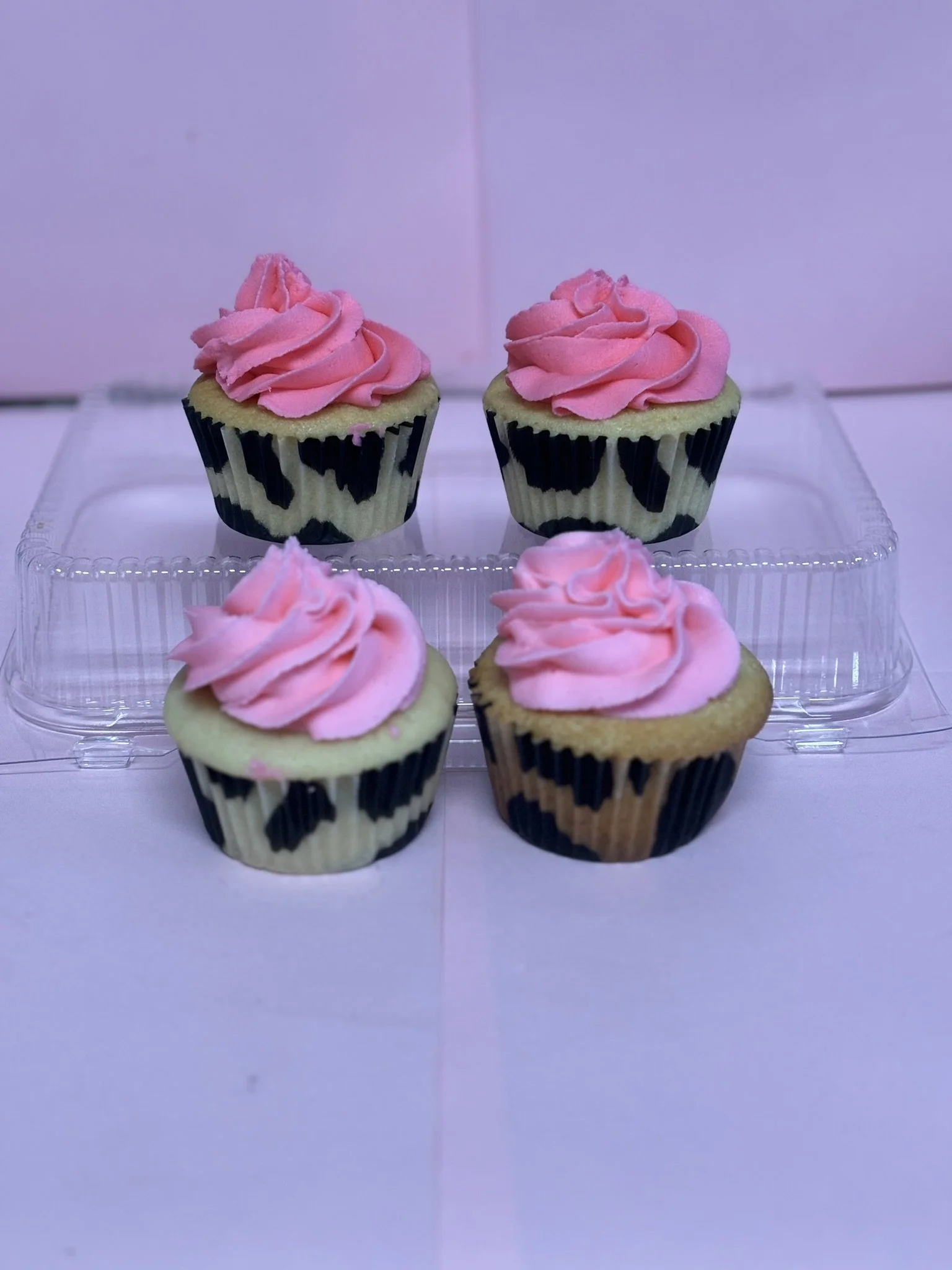 Four cupcakes with pink frosting, in zebra-print cupcake liners, placed on a clear plastic display stand against a pink background.