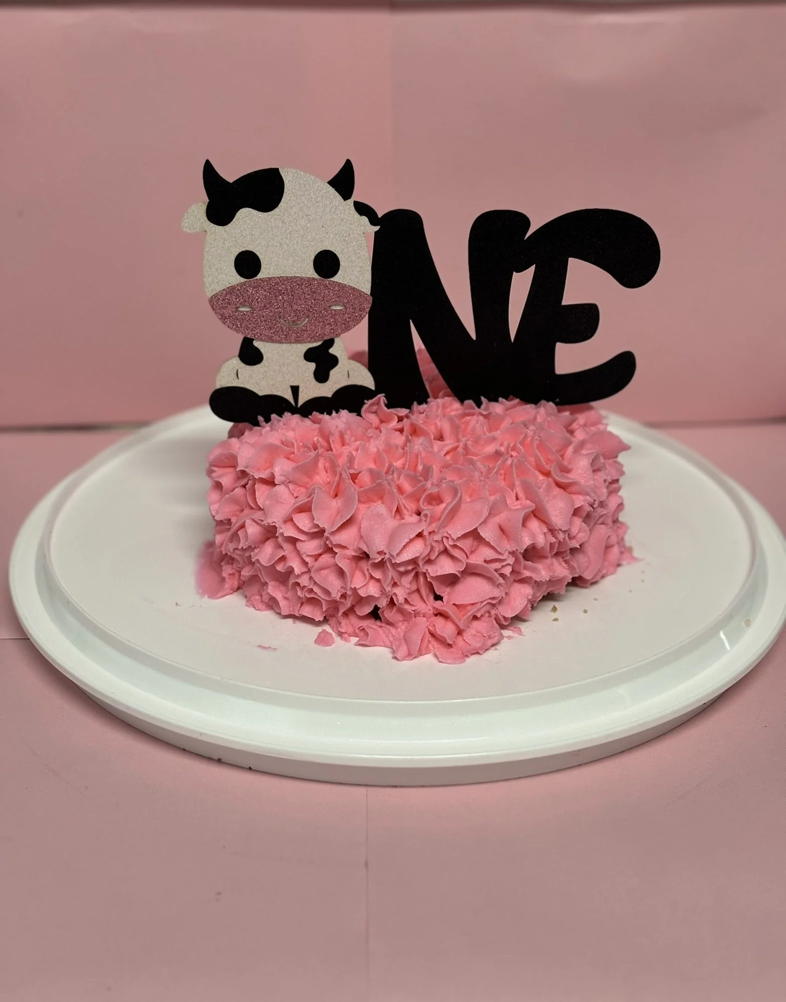 A pink heart-shaped cake decorated with ruffled pink frosting, topped with a black 'LOVE' sign and a cute cow figure with a pink bowtie and glittery pink face on a white plate against a pink background.