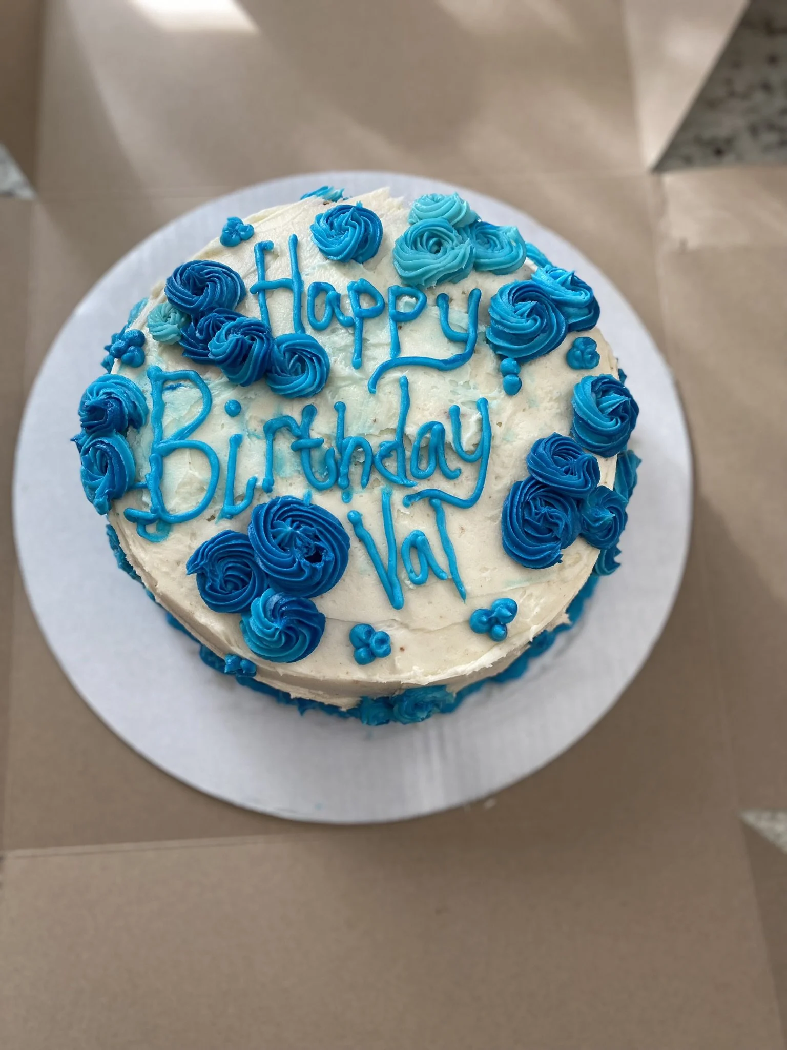 Round birthday cake decorated with blue and turquoise frosting roses, with the message "Happy Birthday Val" written in blue icing on top.