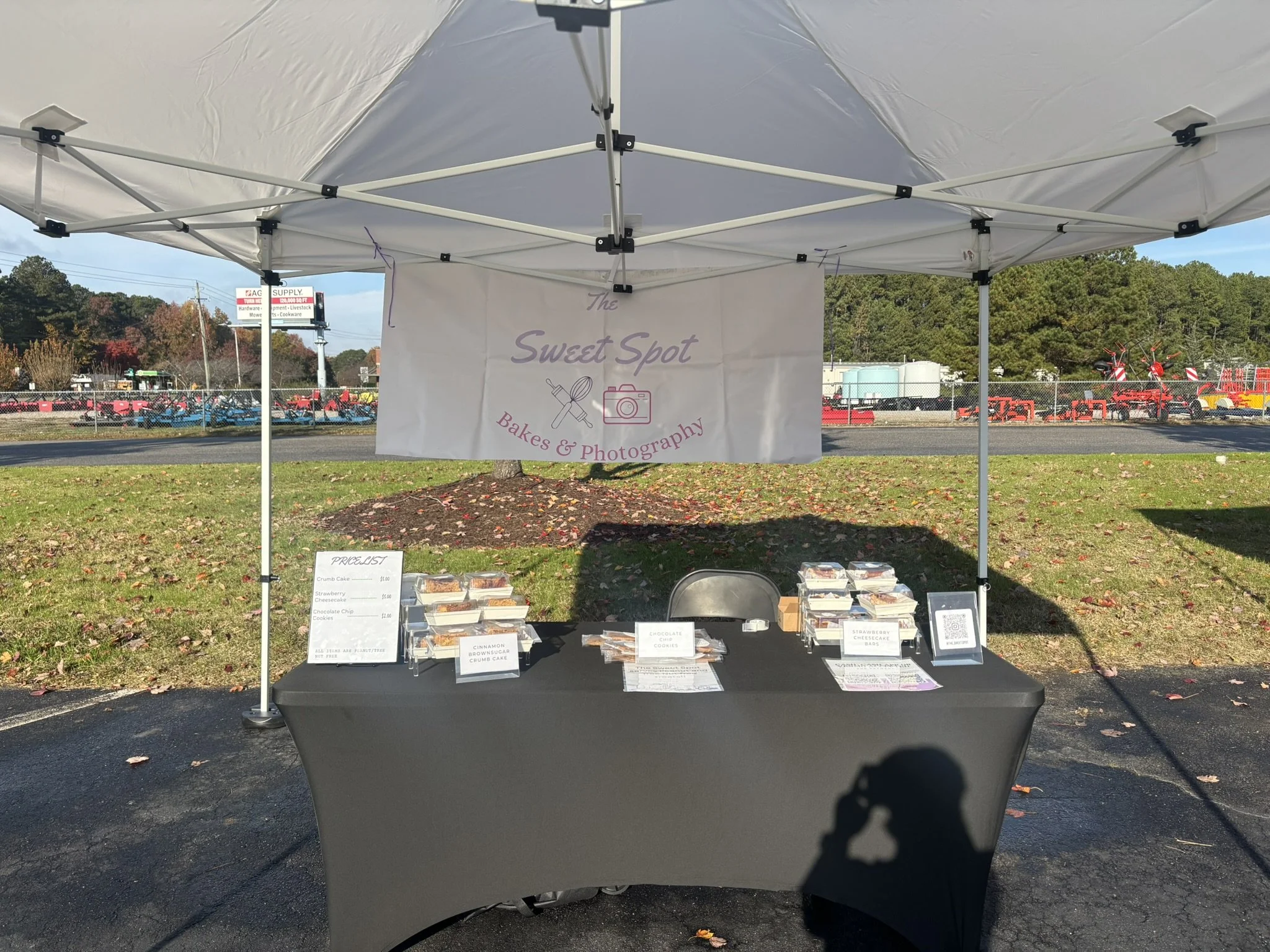 Outdoor market stall for The Sweet Spot Bakes & Photography with baked goods on black table and a white sign hanging from the tent, featuring the shop name, logo, and services.