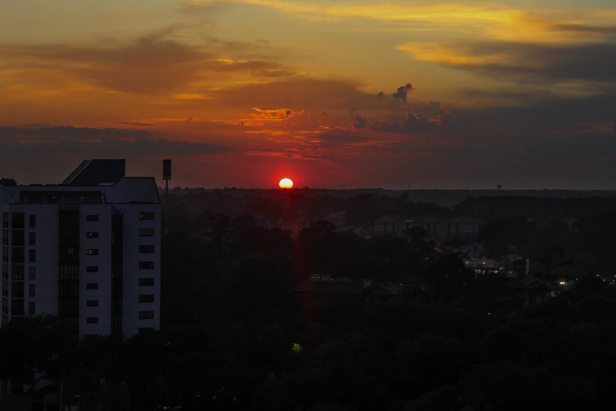 Sunset over a cityscape with silhouettes of buildings and trees, a vibrant orange and yellow sky with scattered clouds.