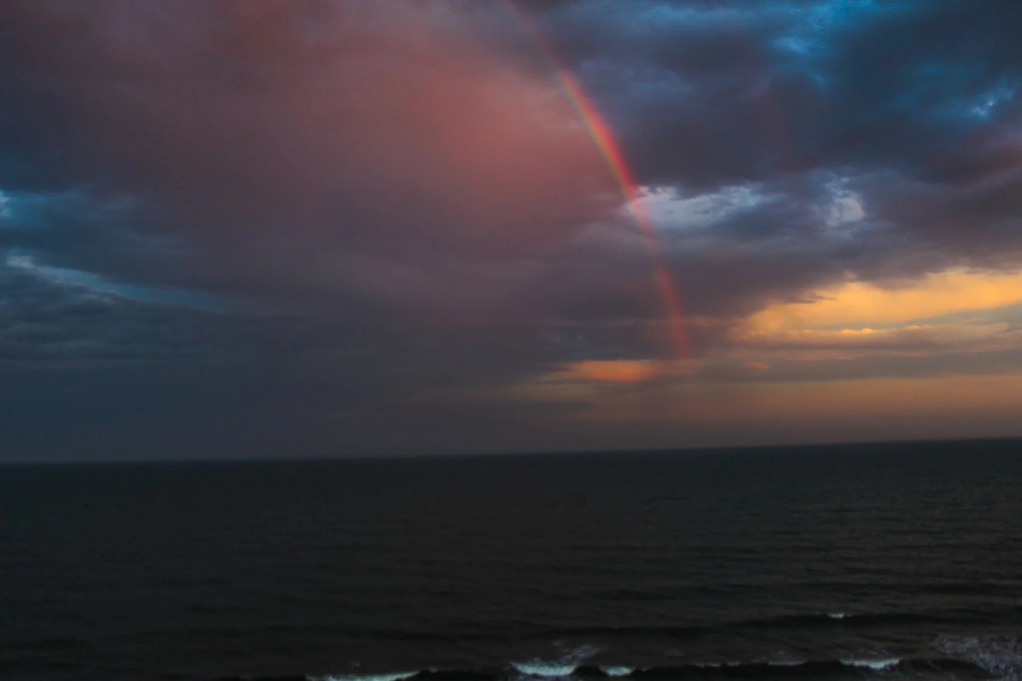 A sunset over the ocean with a dark, cloudy sky and a visible rainbow arching through the clouds.
