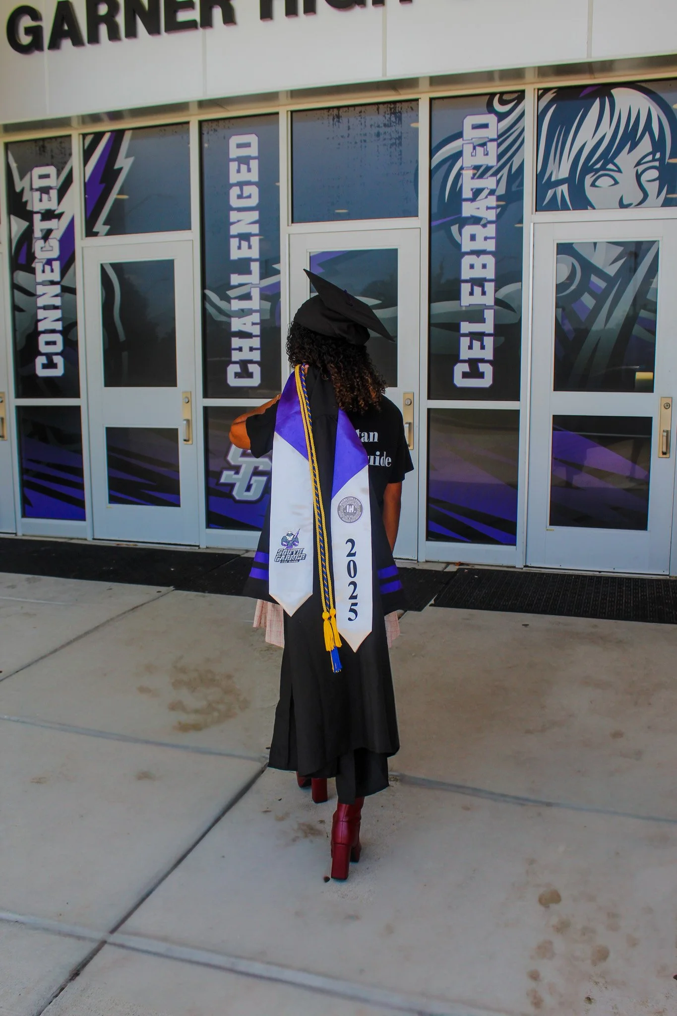 A graduate dressed in black cap and gown with a blue and white stole that reads '2025' , standing outside a building with large windows and doors. The windows display words like 'Connected,' 'Challenged,' 'Celebrated,' and have a graphic of a person'