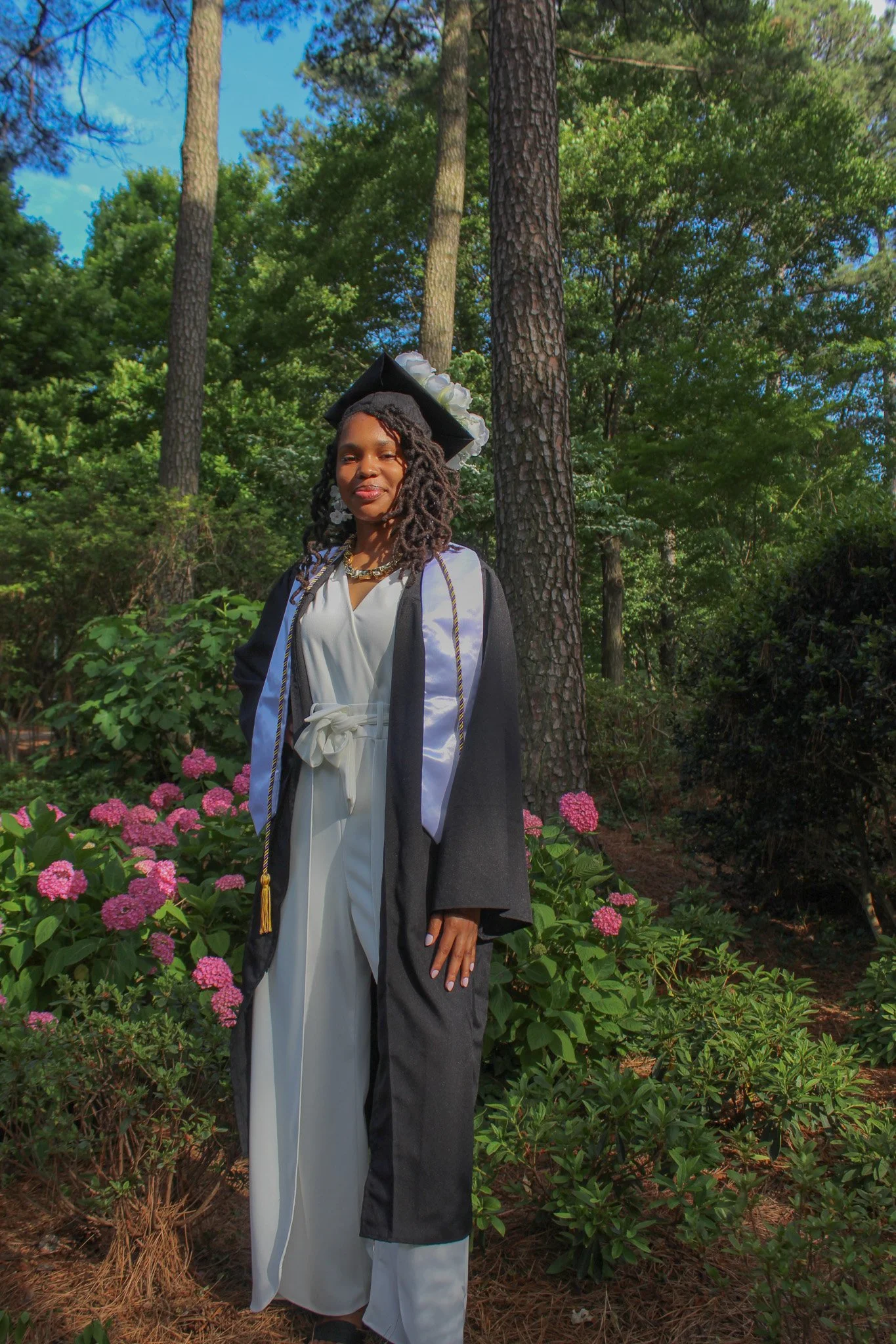 A woman in graduation attire standing outdoors among pink flowers and green trees.