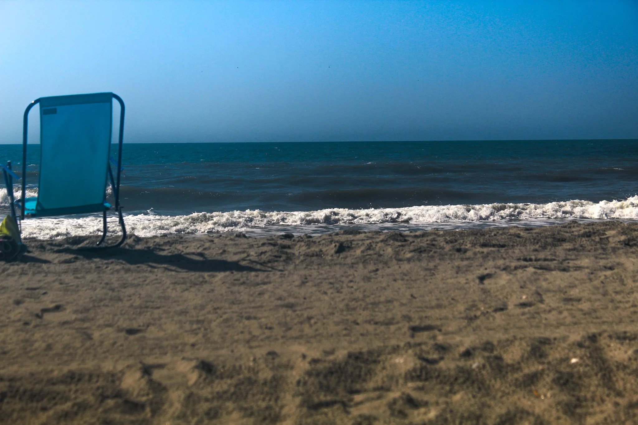 Empty blue beach chair on sandy shore facing the ocean, with small waves and a clear blue sky.