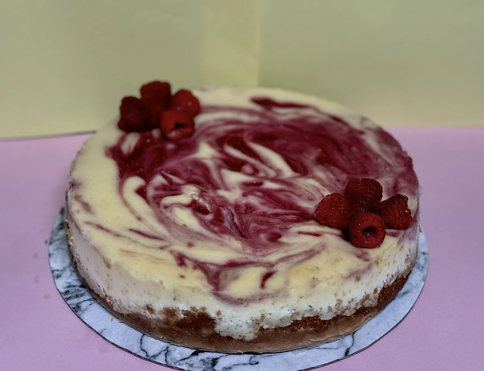 Cheesecake with raspberry swirl pattern and fresh raspberries on top, on a pink surface with a pastel yellow background.