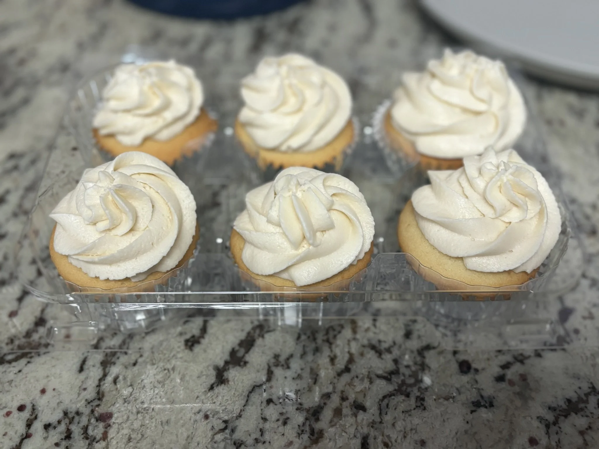 Six cupcakes with white frosting in a plastic tray on a granite countertop.