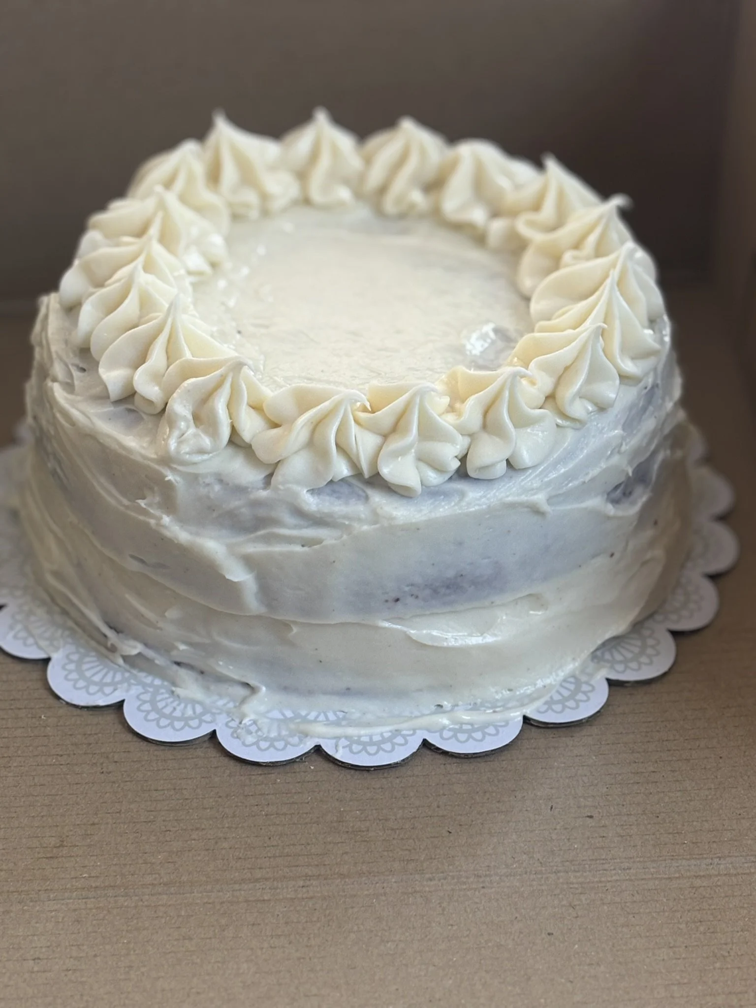 A round, white frosted cake decorated with piped white frosting around the top edge, resting on a doily