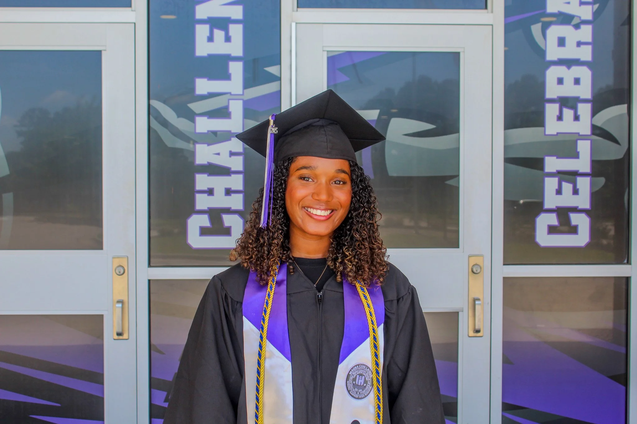 A young woman in a graduation cap and gown standing in front of glass doors with purple 'CHALLENGE CENTER' signs, smiling at the camera.