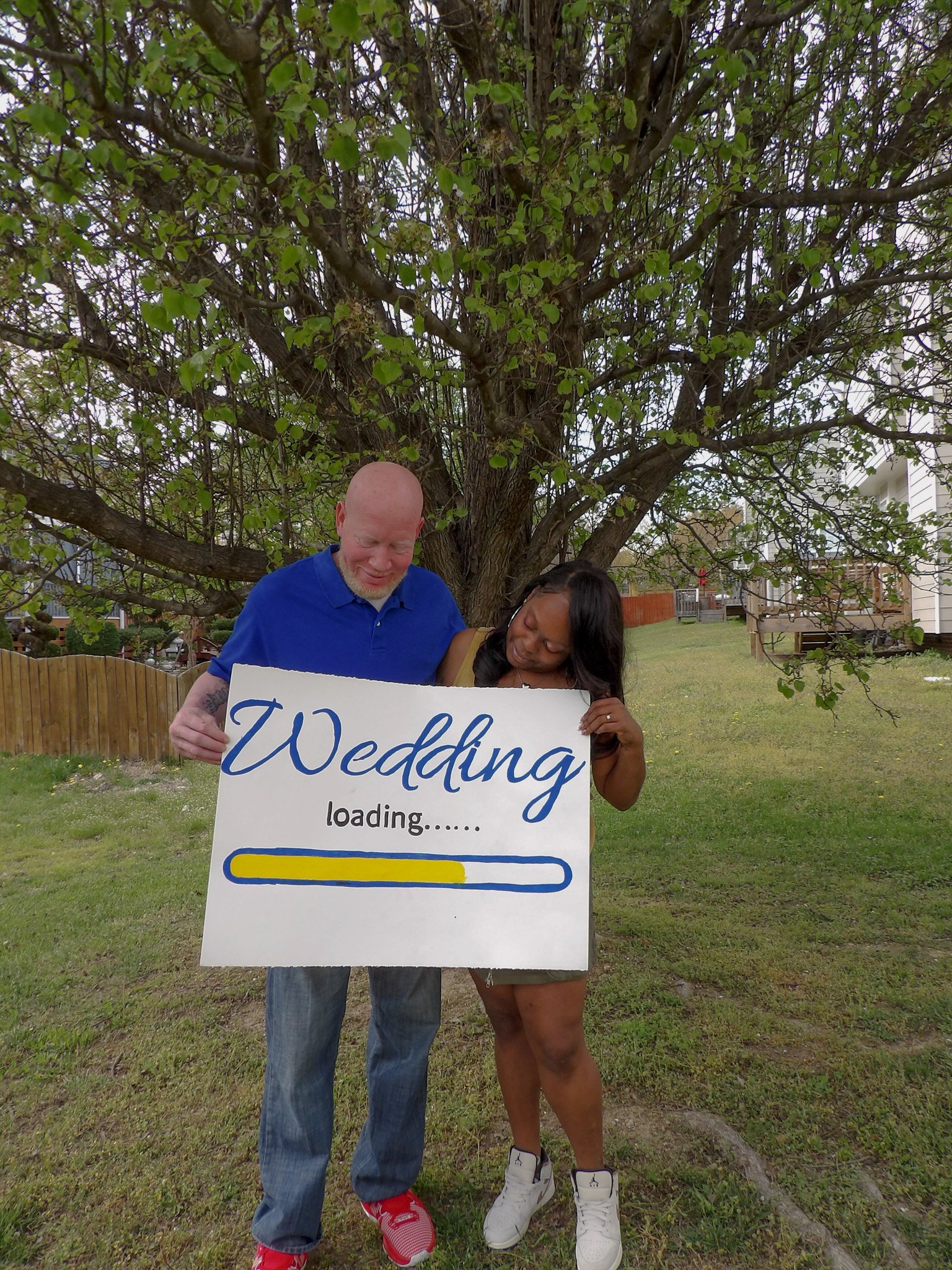 A couple standing outdoors under a large tree, holding a sign that says "Wedding loading....." with a loading bar partly filled.