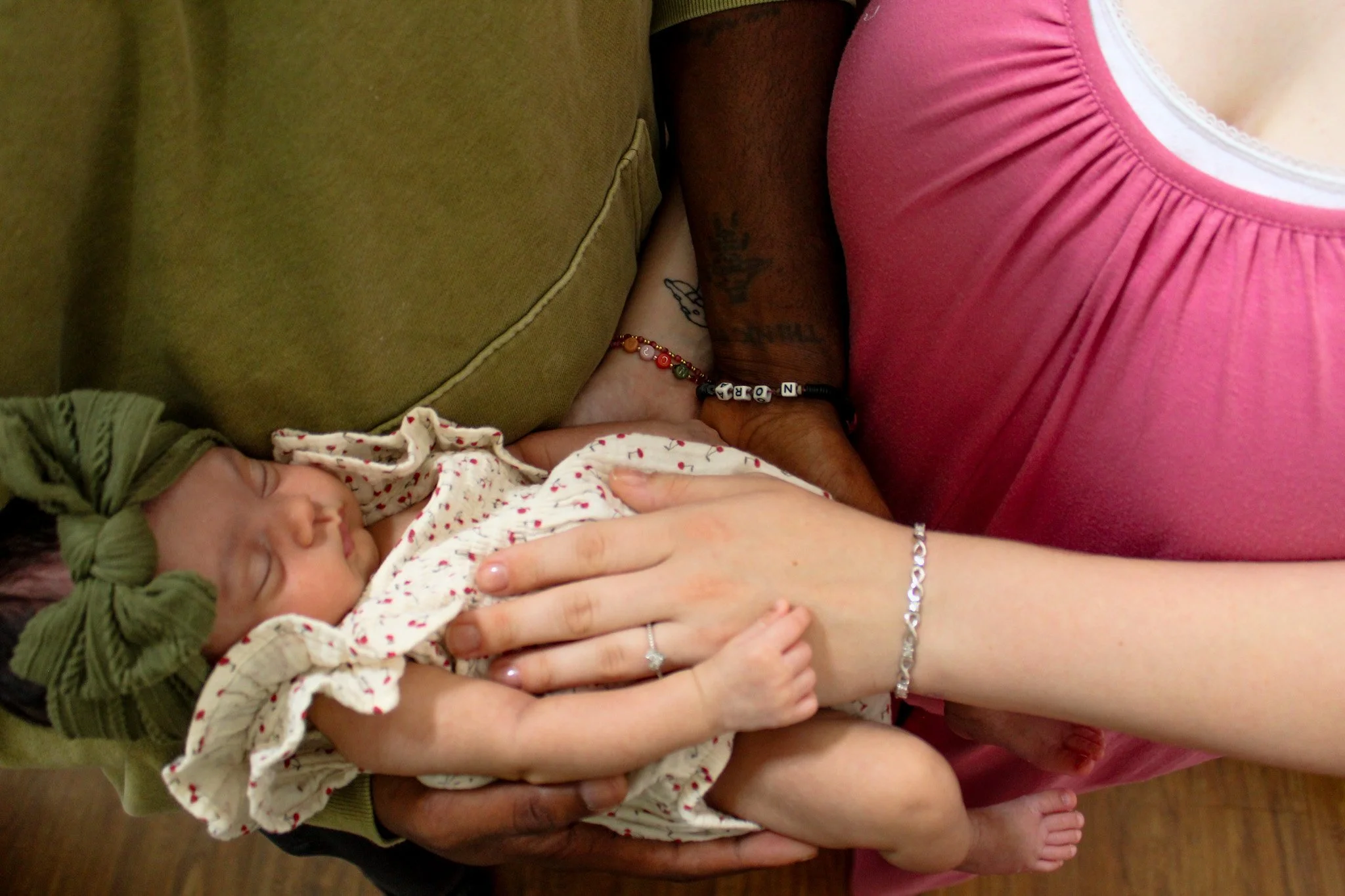 Close-up of a couple holding a sleeping baby girl dressed in a white onesie with red hearts, wearing a green bow headband.