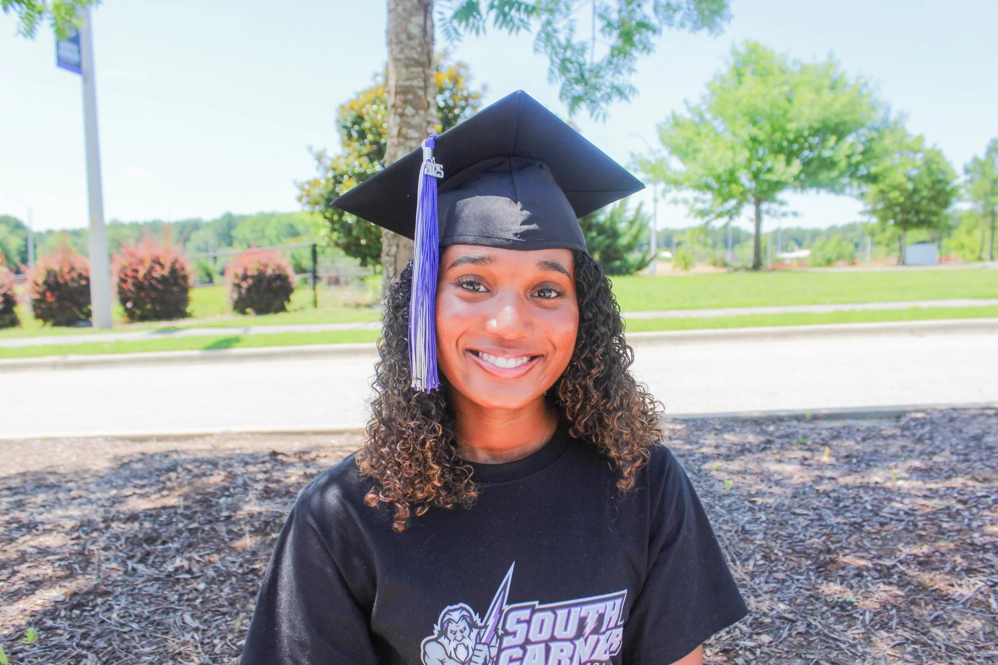 Young woman smiling outdoors wearing a black graduation cap and a black T-shirt with a graphic and text.