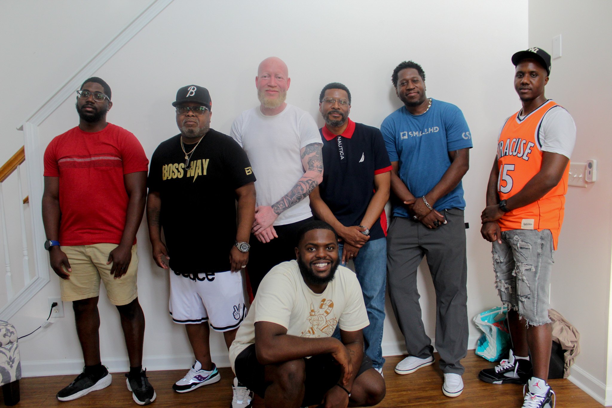 A group of eight men posing indoors, standing against a white wall, with one man kneeling in front.