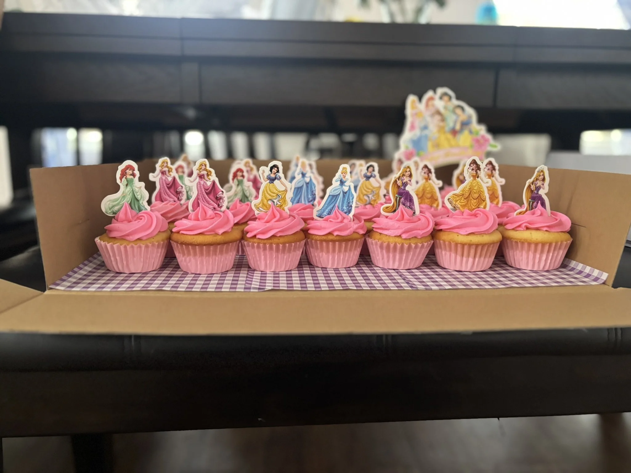 Cupcakes with pink frosting and princess toppers, arranged in a tray with purple checkered paper.