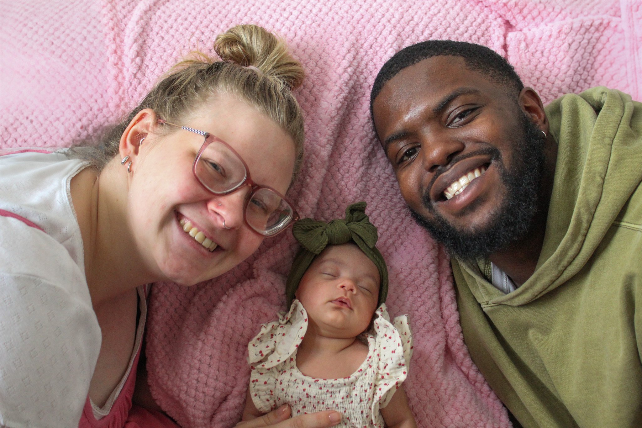 A smiling woman with glasses and earrings, a sleeping baby with a green headband, and a smiling man lying on a pink blanket, all taking a selfie together.